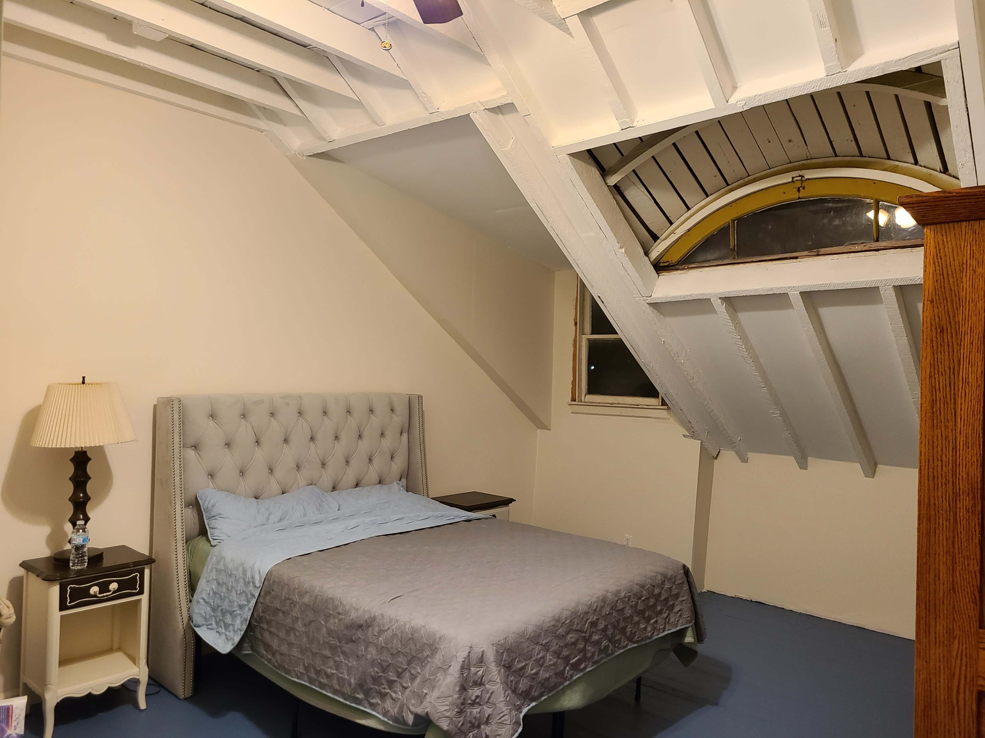A sparsely furnished bedroom with a tufted bed frame, a nightstand, and exposed rafters in the ceiling.