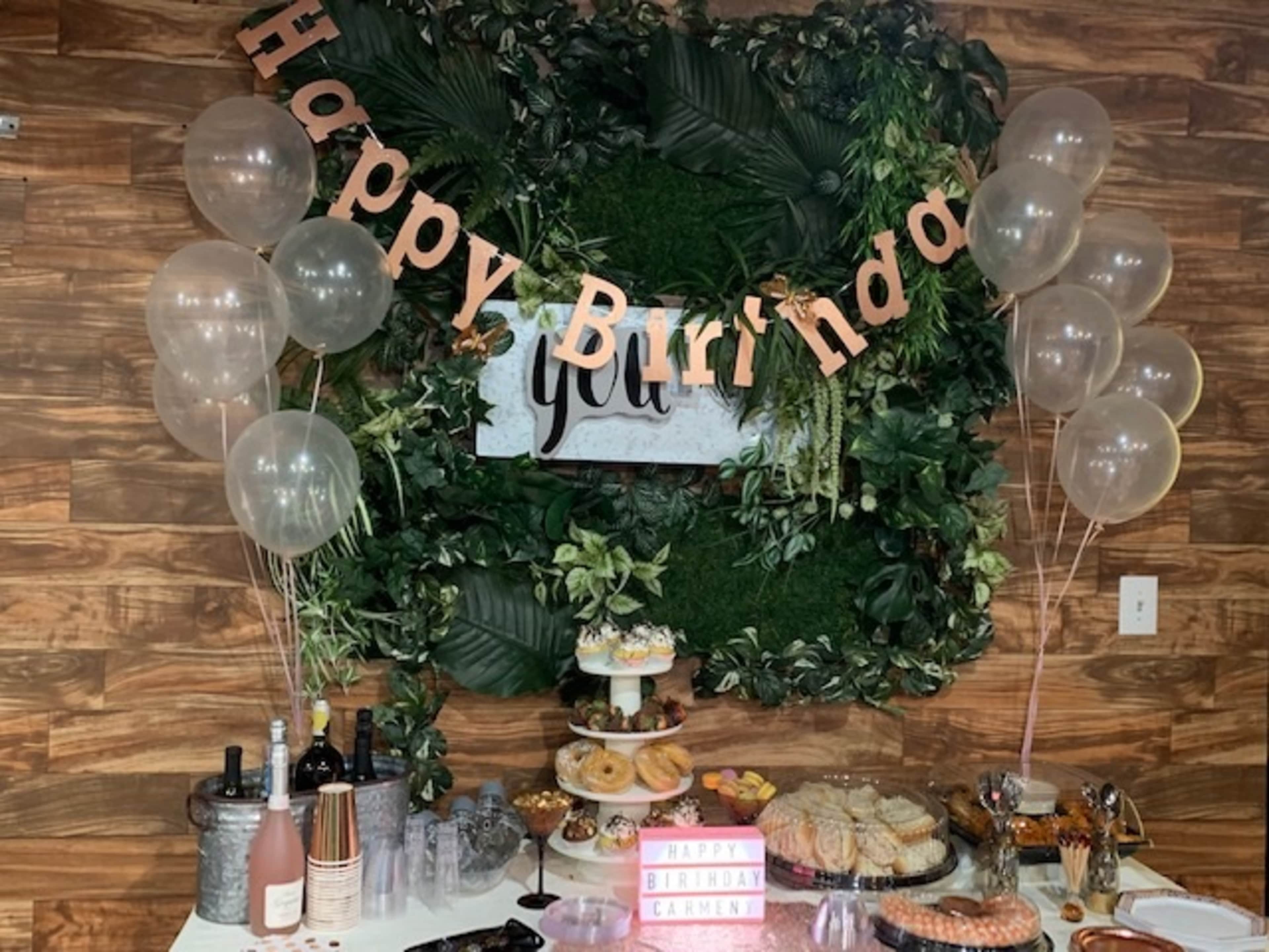 A birthday celebration setup featuring a table with various snacks and drinks, a "Happy Birthday" banner, and balloons against a green foliage backdrop.