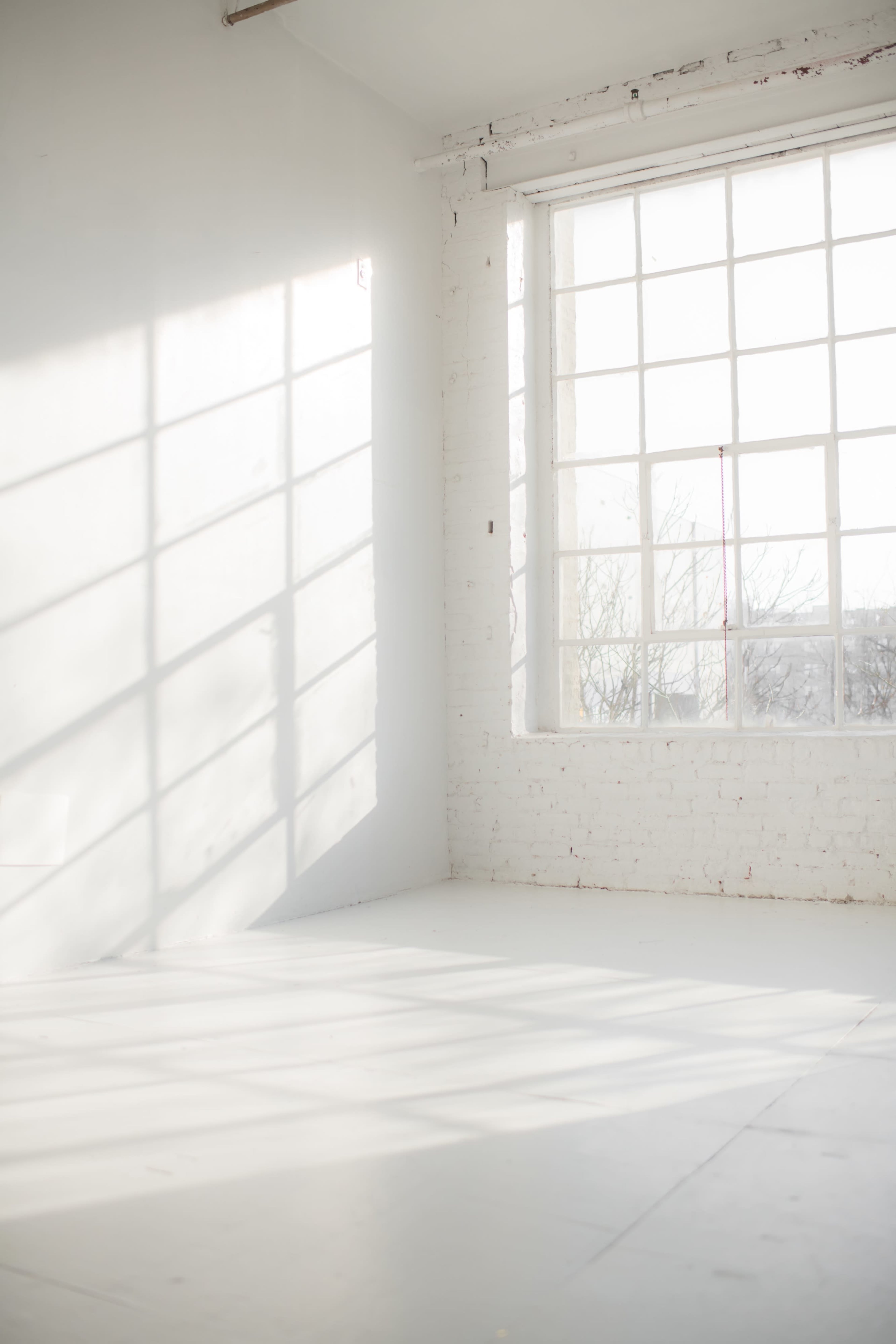 A bright, empty room features a large window casting shadows on the white walls and floor.