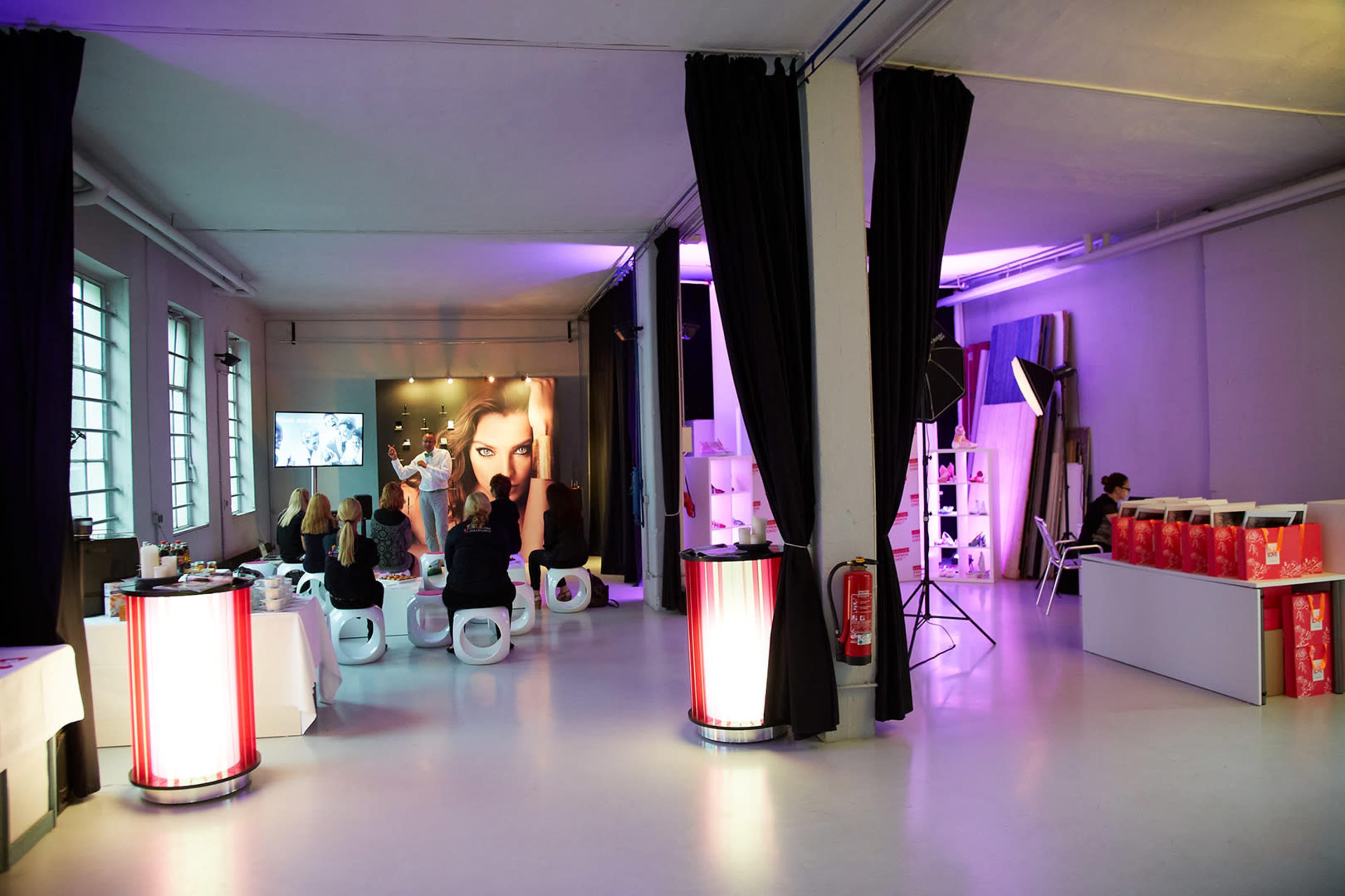 The image shows a modern event space with guests seated in white chairs, a screen displaying a woman’s face, and product displays in the background.