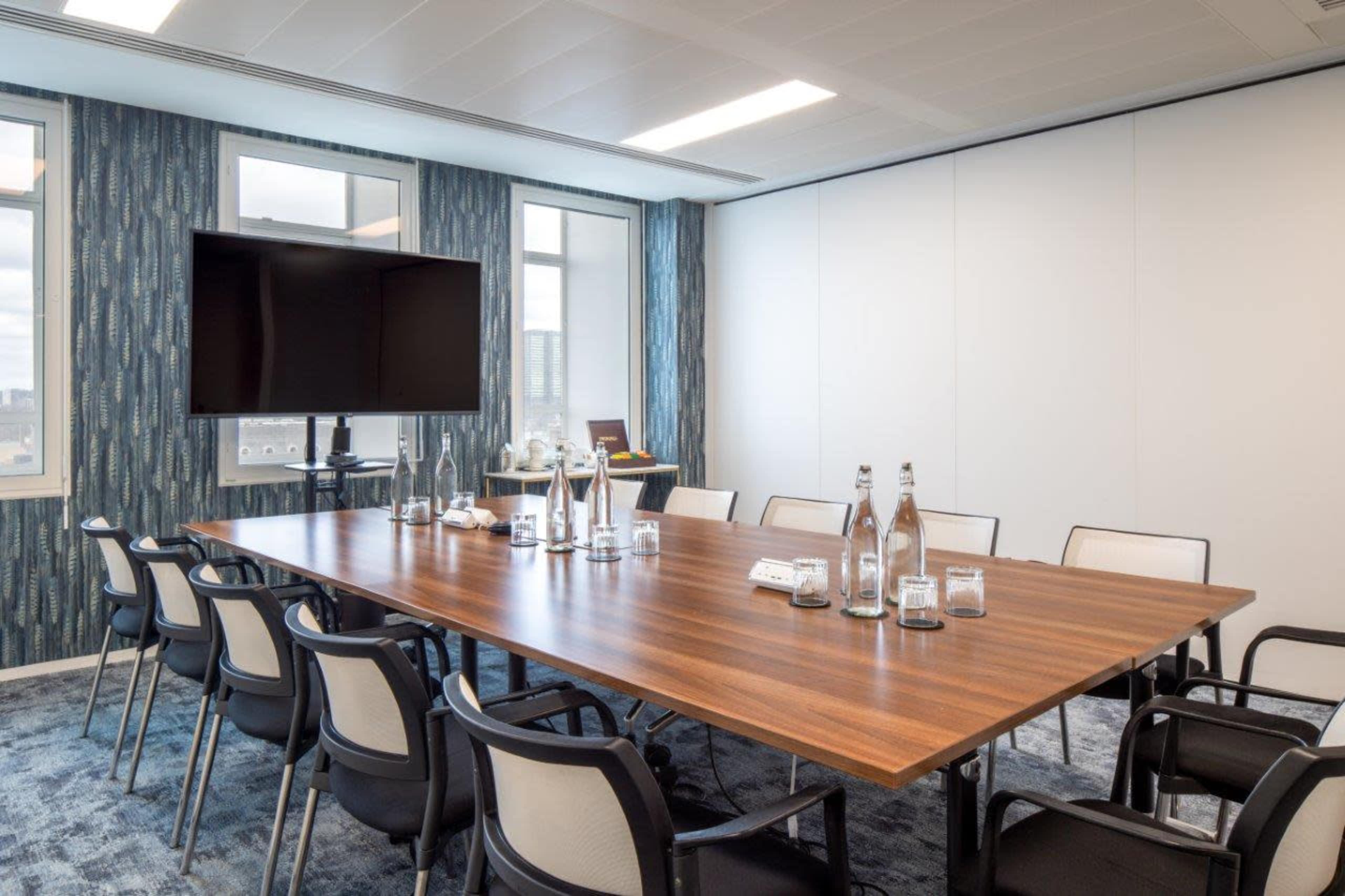 A modern conference room features a large wooden table surrounded by black chairs, equipped with a television for presentations and large windows letting in natural light.
