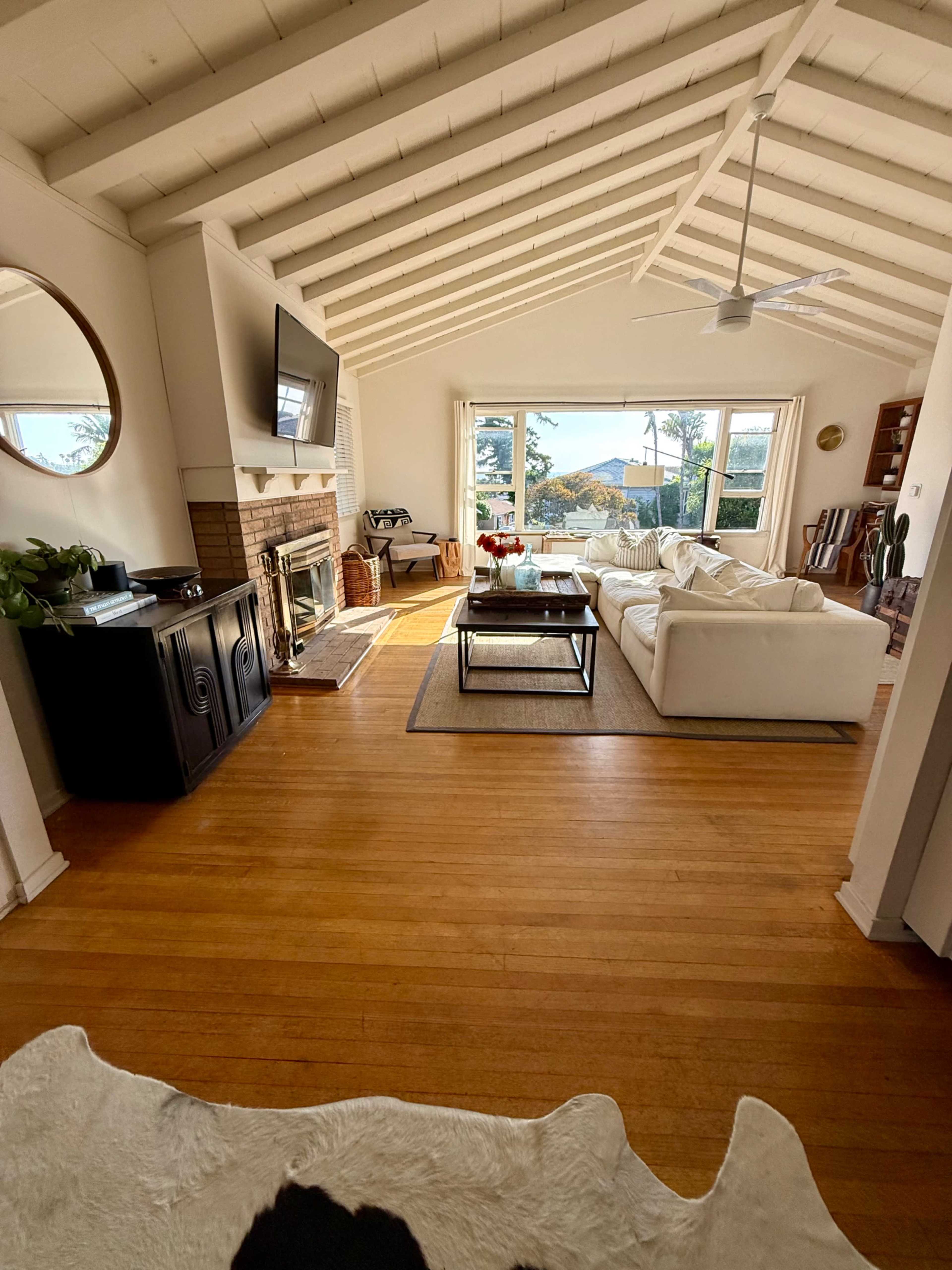 The image shows a spacious living room with a white sofa, a coffee table, a fireplace, and large windows that overlook an outdoor view.