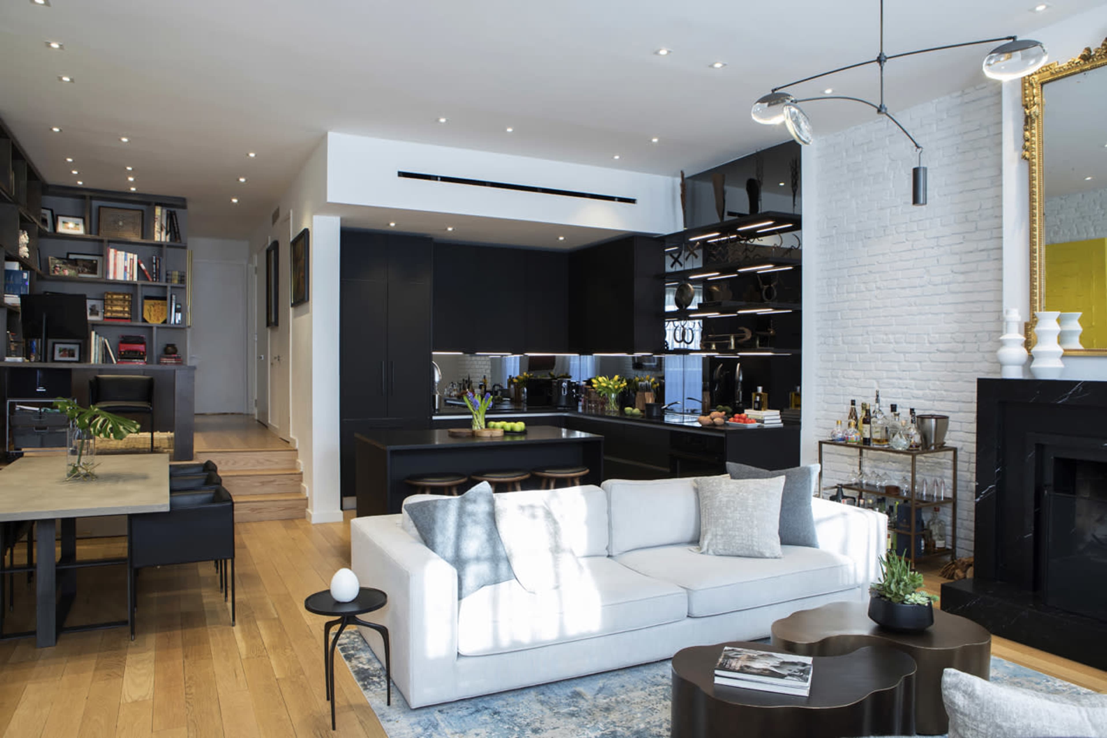 The image shows a modern open-concept living area featuring a light-colored sofa, a dark dining table, and a sleek kitchen with black cabinetry.