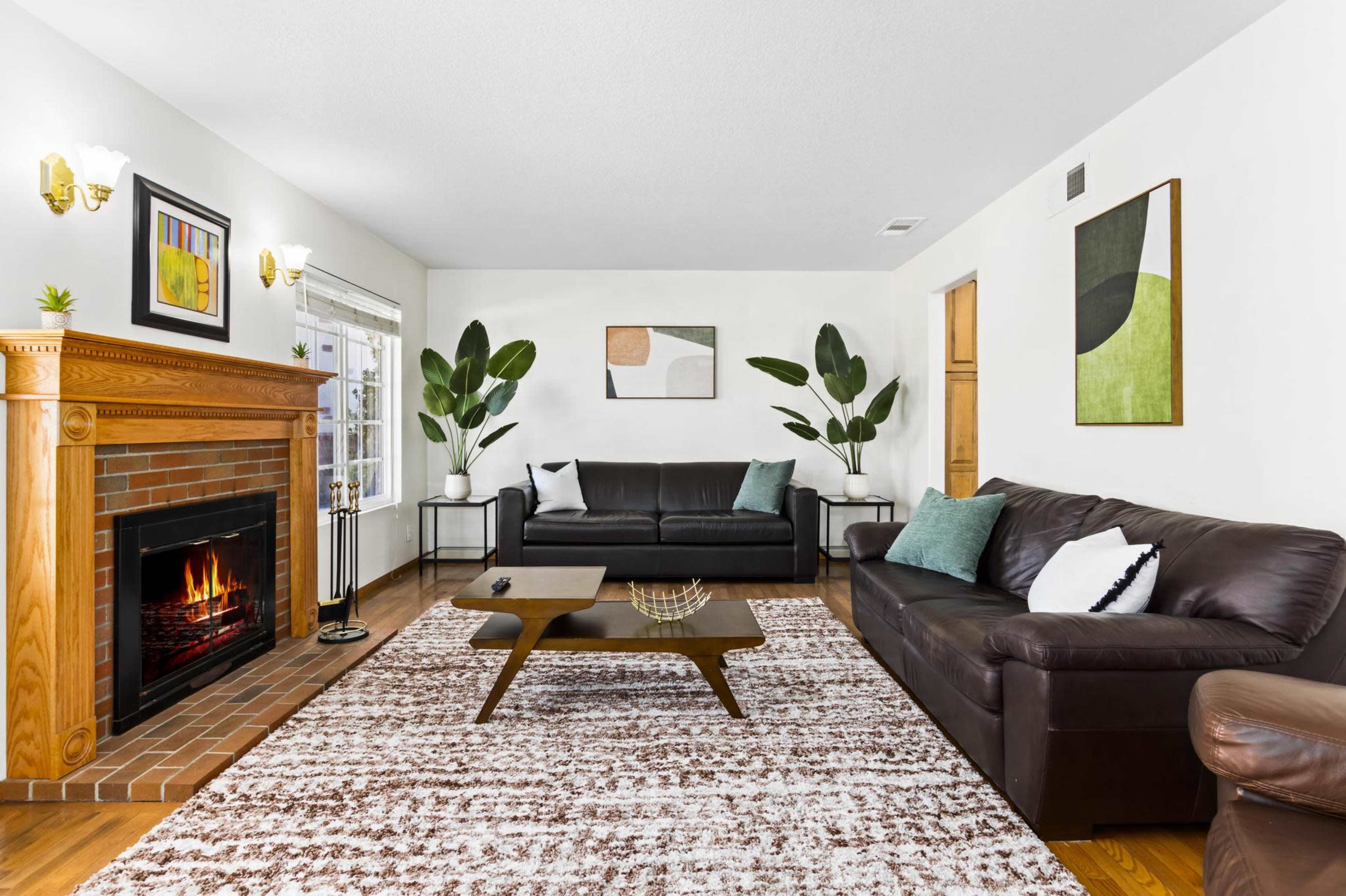 A living room with a brick fireplace, two leather sofas, a coffee table, and plants beside large windows.