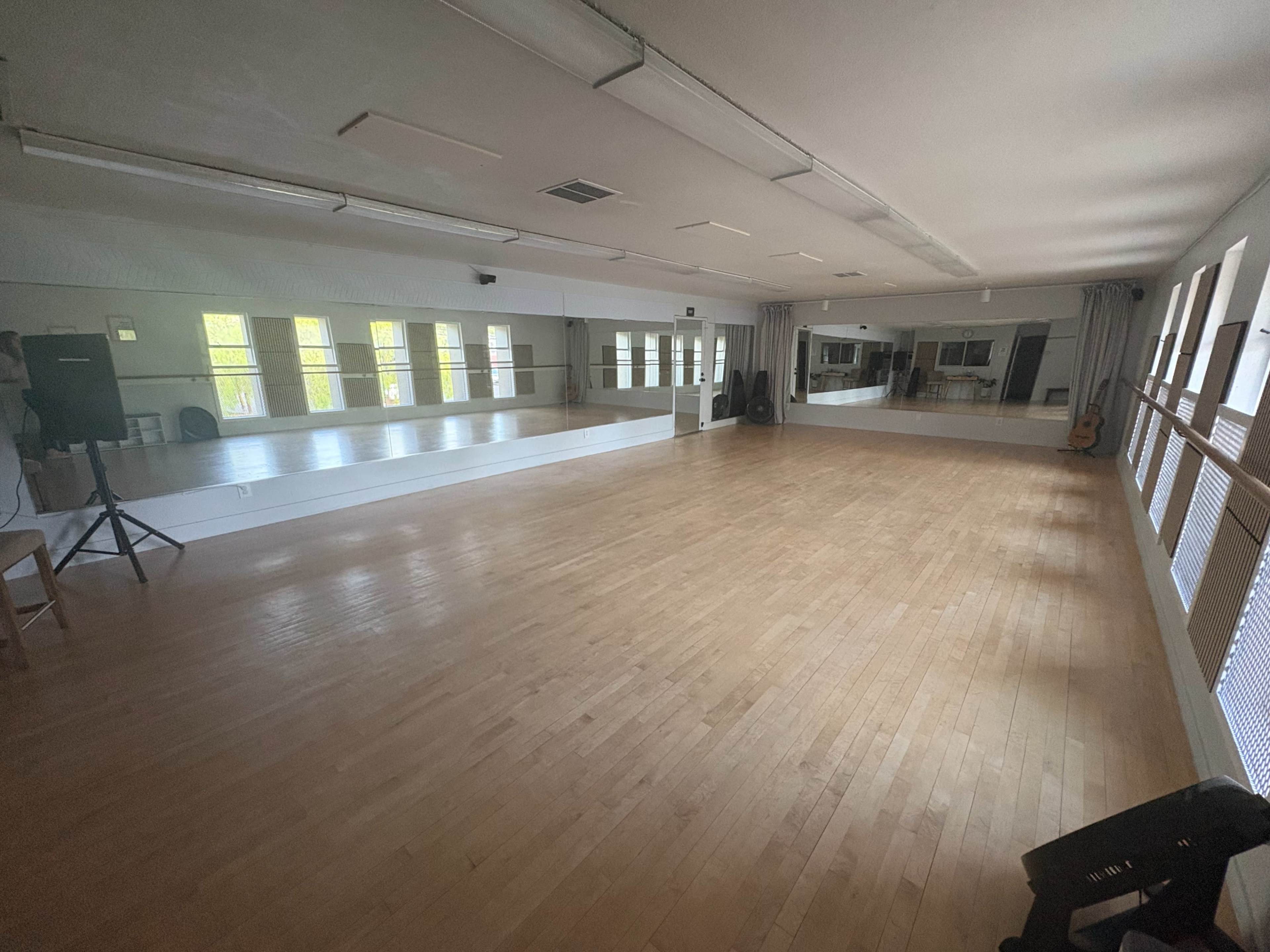 The image depicts a spacious dance studio with wooden floors, large mirrors on one wall, and natural light coming through windows.