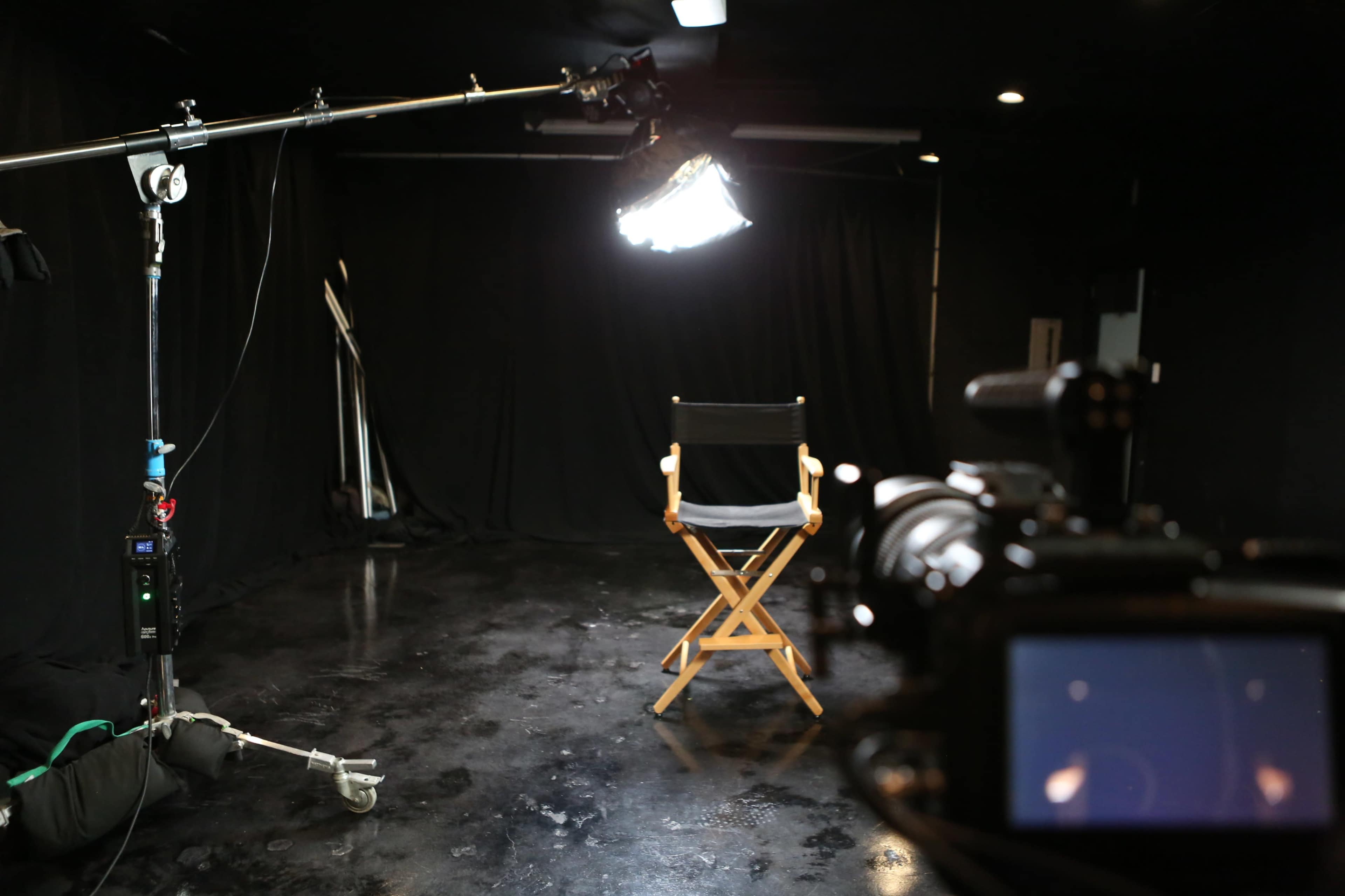 A film studio setup with a wooden director's chair facing a camera, illuminated by a large light source against a backdrop of black curtains.
