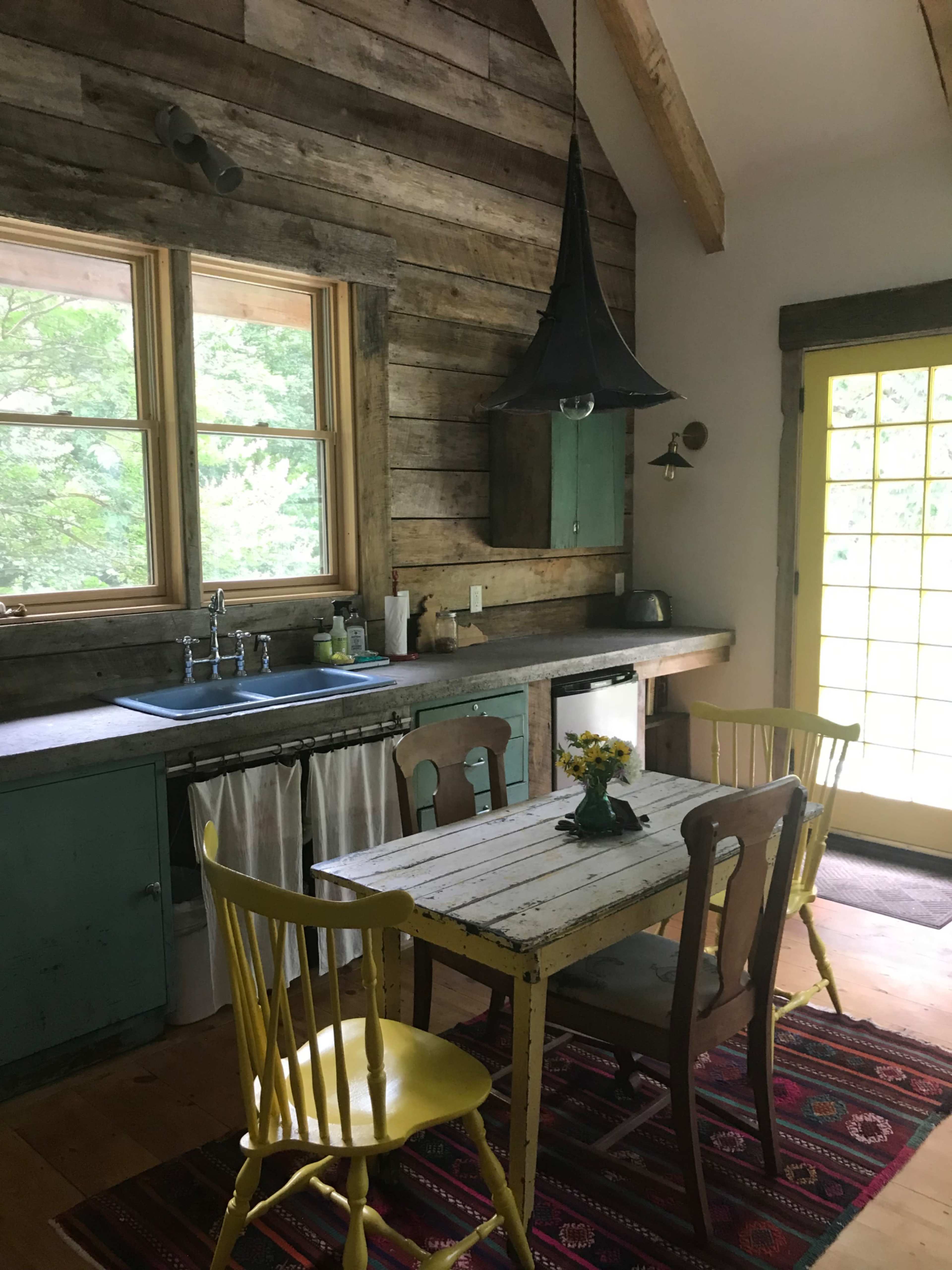 A cozy kitchen features a wooden table surrounded by yellow and brown chairs, with a rustic countertop and a bright yellow door leading outside.