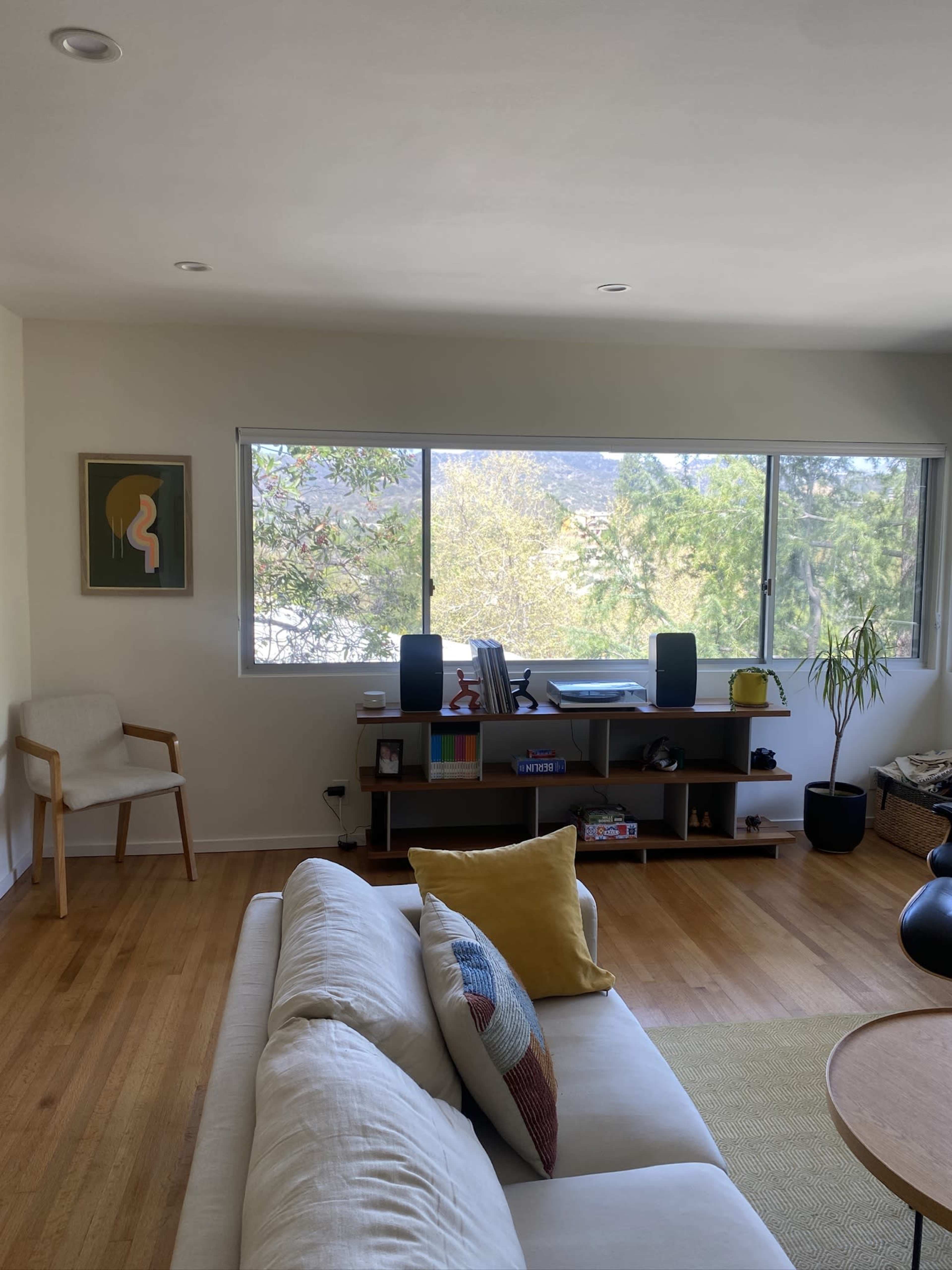 The image shows a modern living room with a white couch, a wooden shelving unit, and large windows overlooking greenery outside.