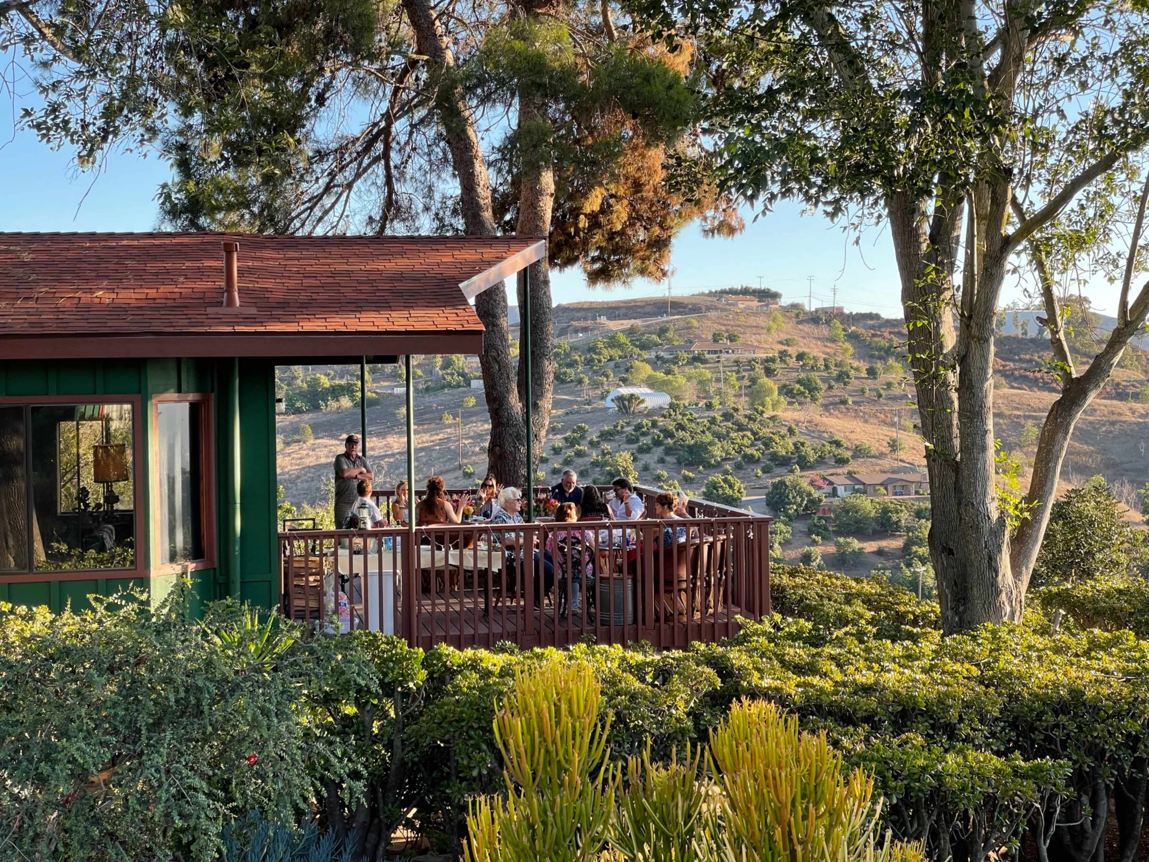 A group of people is gathered around a long table on a patio of a wooden building, surrounded by greenery and rolling hills.