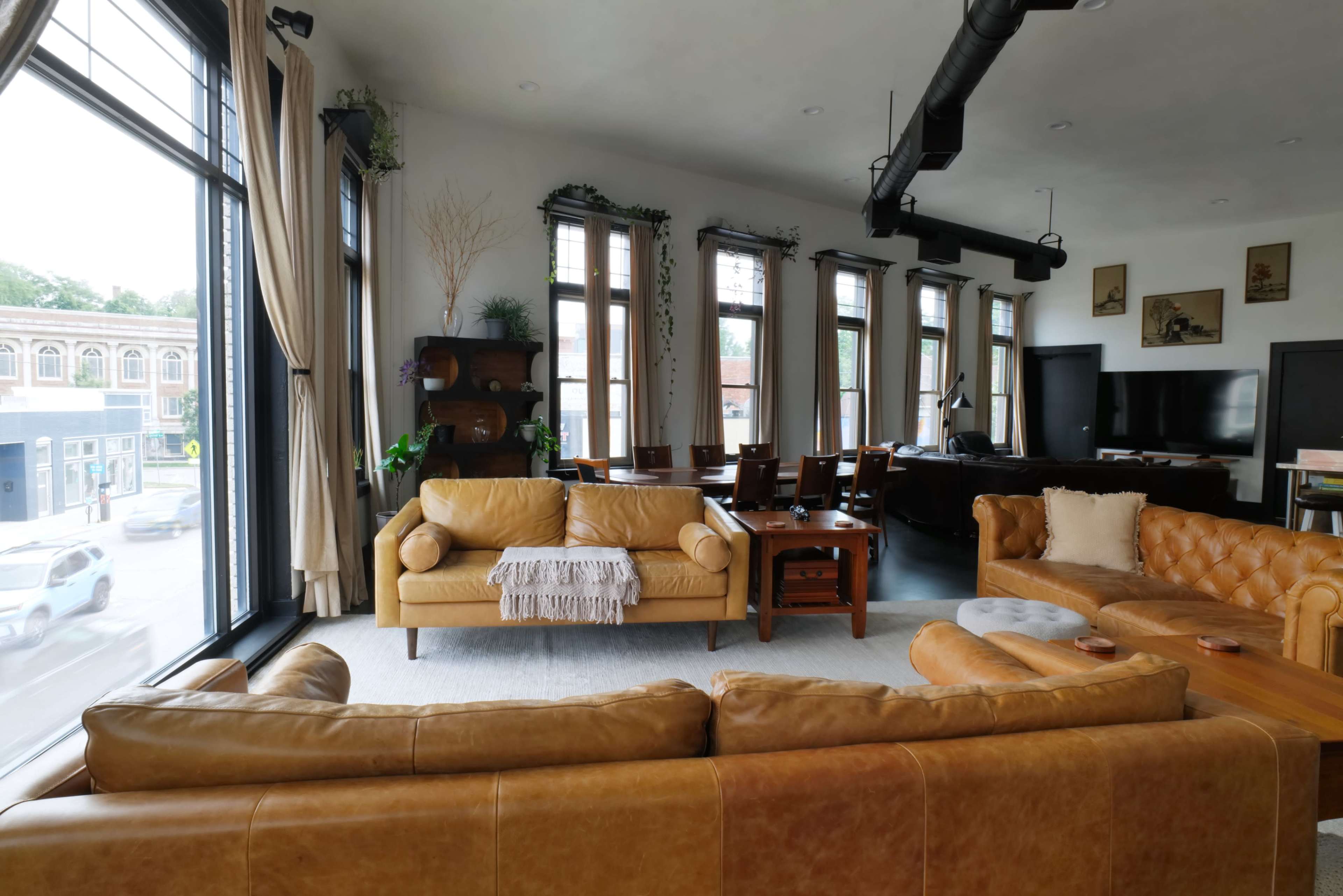The image shows a spacious living area with large windows, featuring a tan leather sectional sofa, wooden furniture, and a dining table in the background.