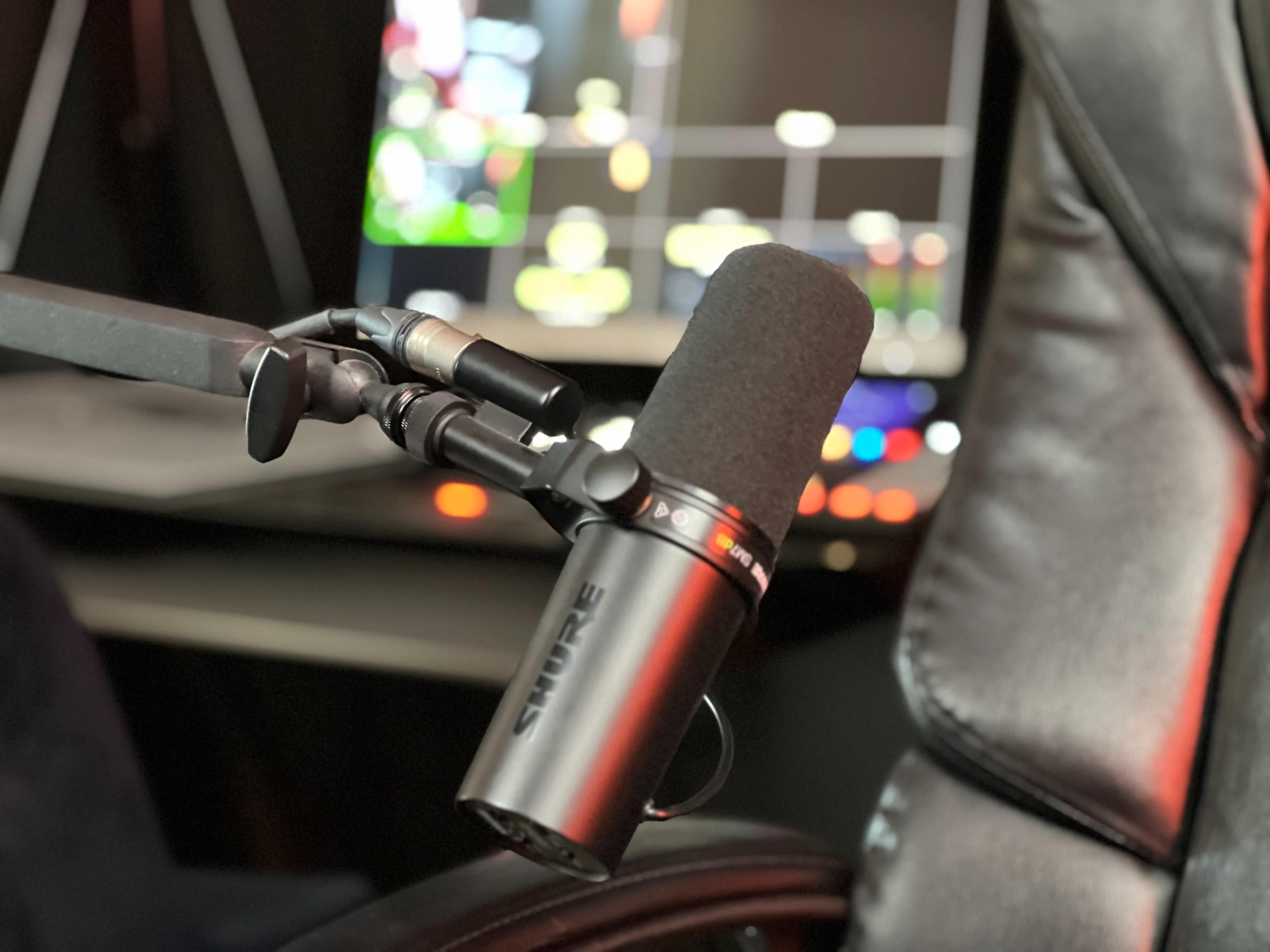 A Shure microphone is attached to an adjustable arm near a computer setup with colorful lights in the background.
