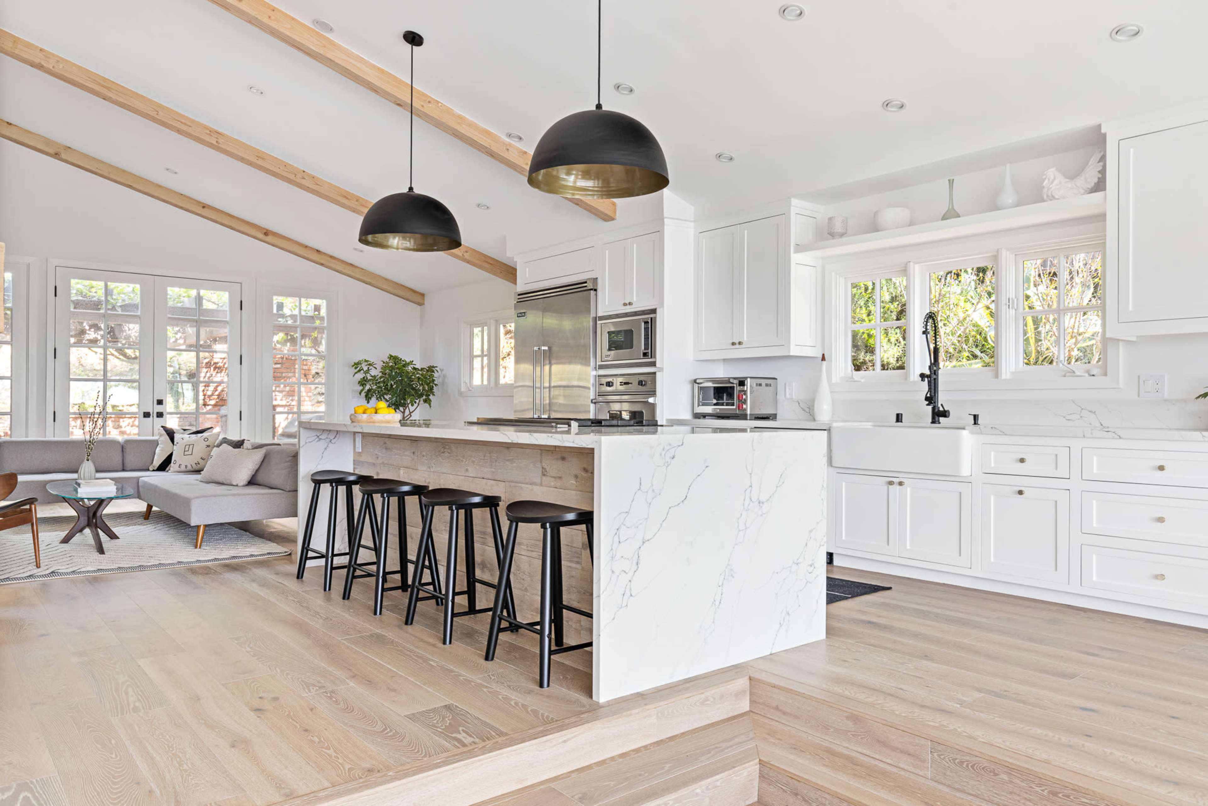 A modern kitchen featuring white cabinetry, a large island with marble countertops, and black bar stools under pendant lighting, adjacent to a bright living area.