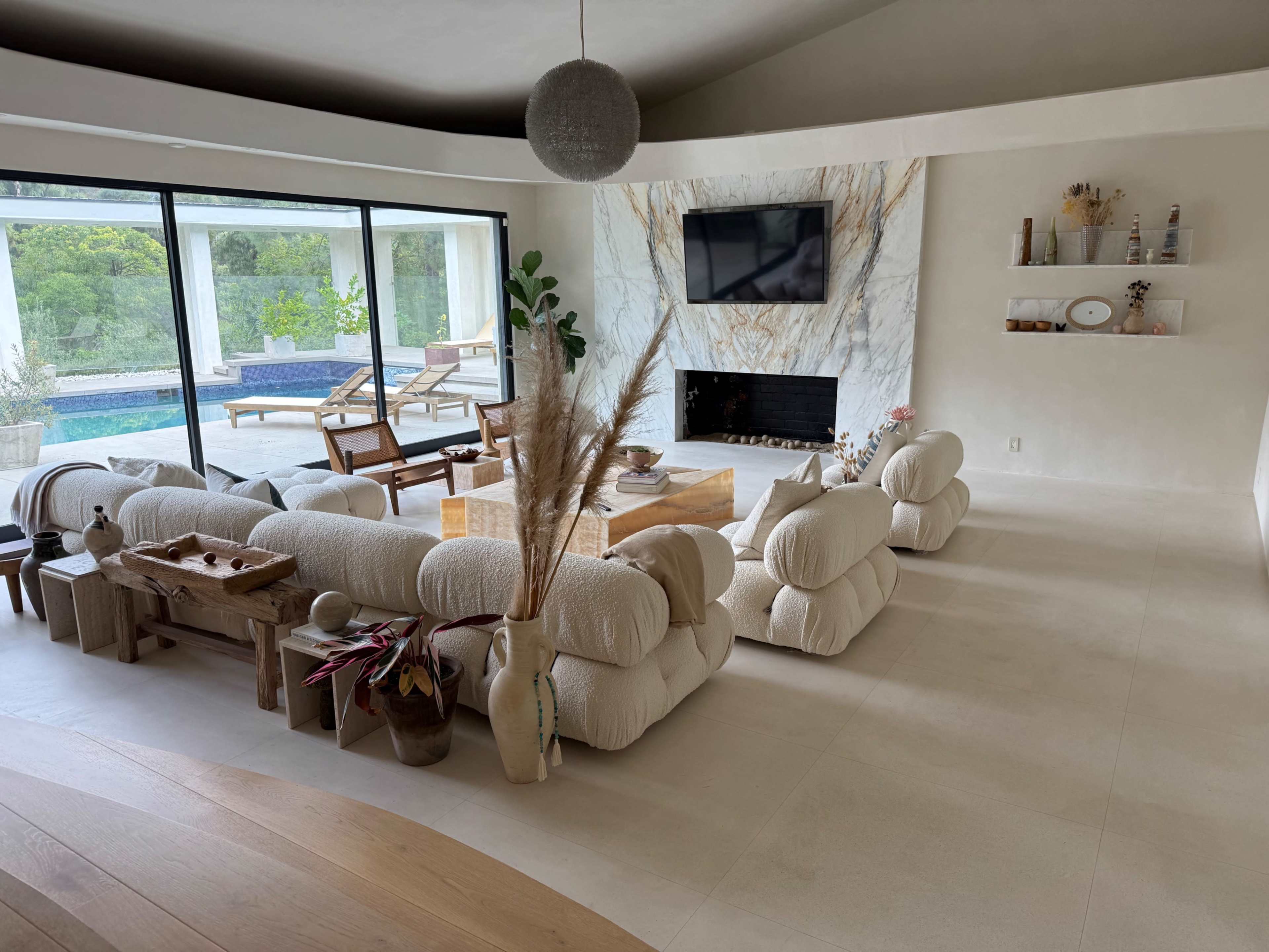 The image shows a modern living room with plush seating, a coffee table, and a wall-mounted television, overlooking a pool through large glass doors.