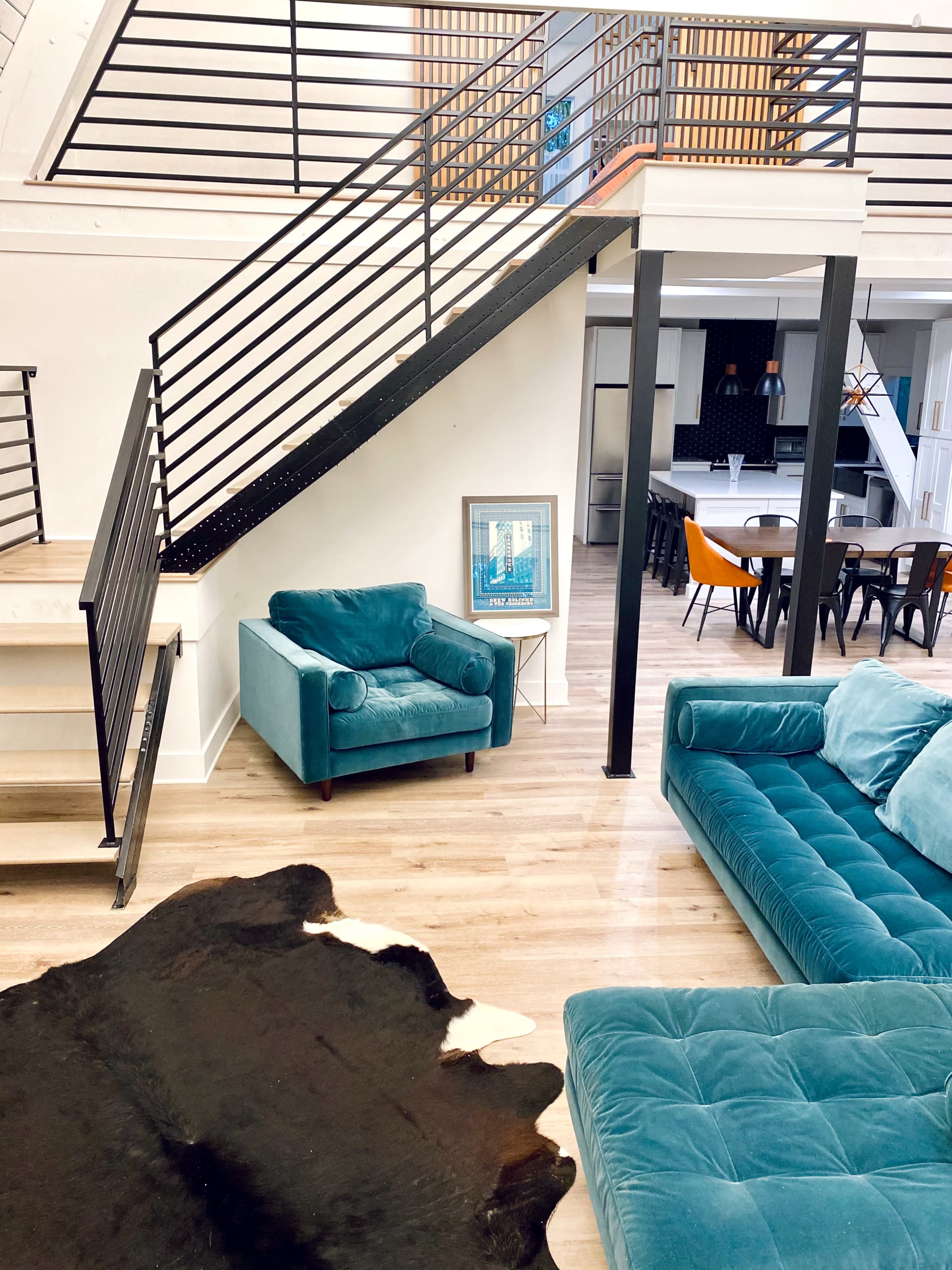 A modern living space featuring a teal couch and armchair, a black and white cowhide rug, a staircase with a metal railing, and a dining area in the background.