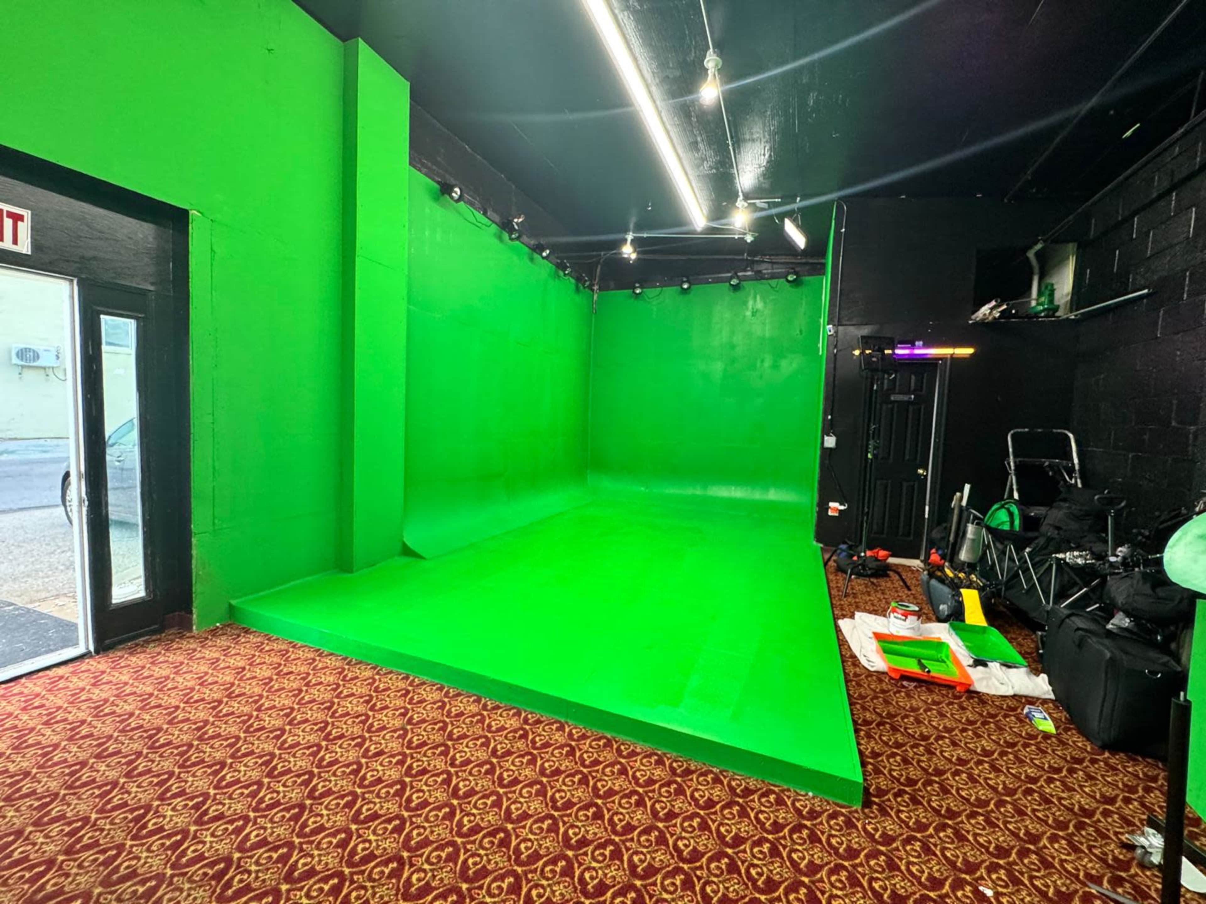 The image shows an indoor space featuring a green screen backdrop, illuminated by overhead lights, with a patterned carpet and some equipment scattered on the floor.