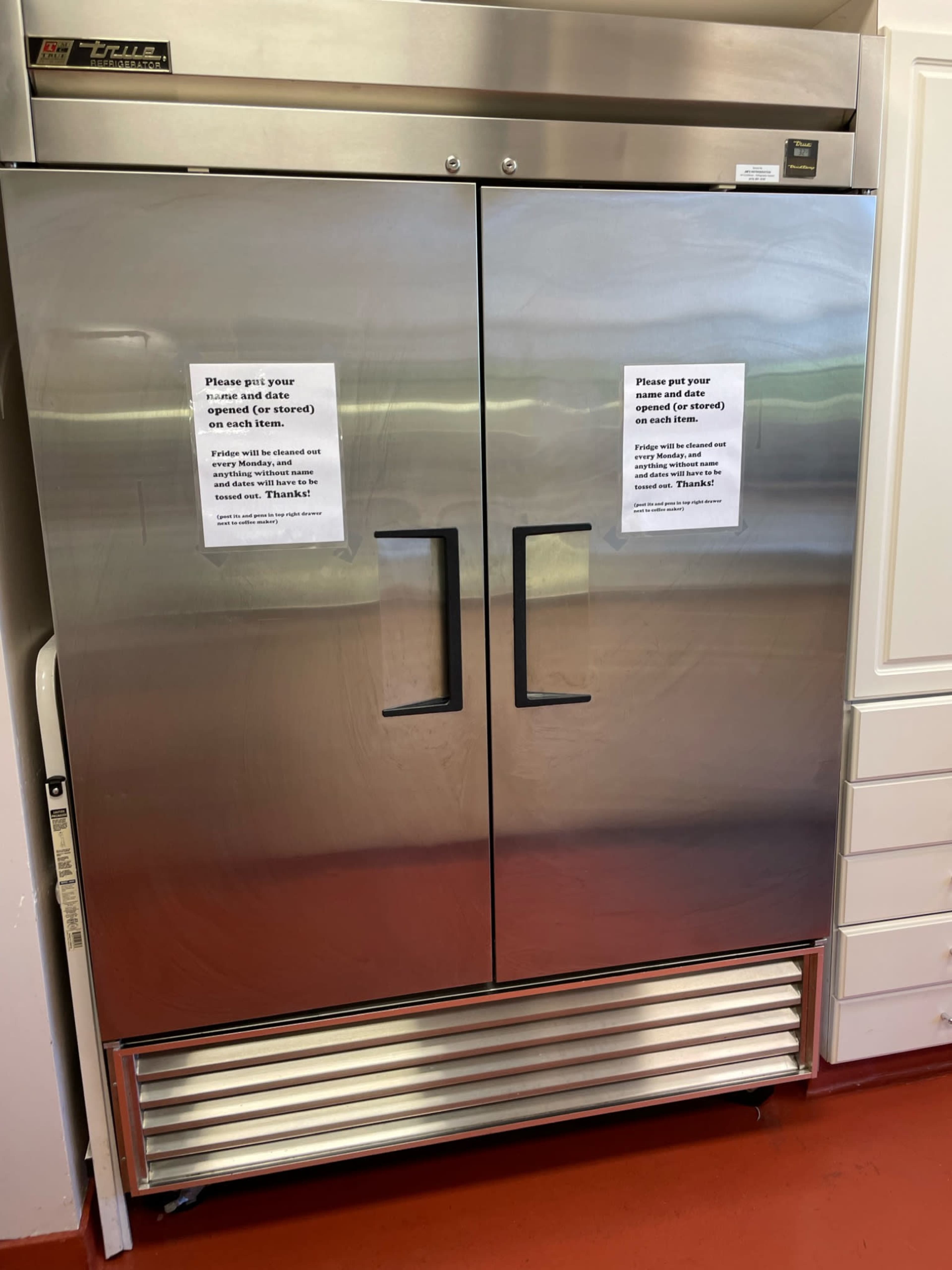 A stainless steel commercial refrigerator with two doors displays two notice signs taped to the front.