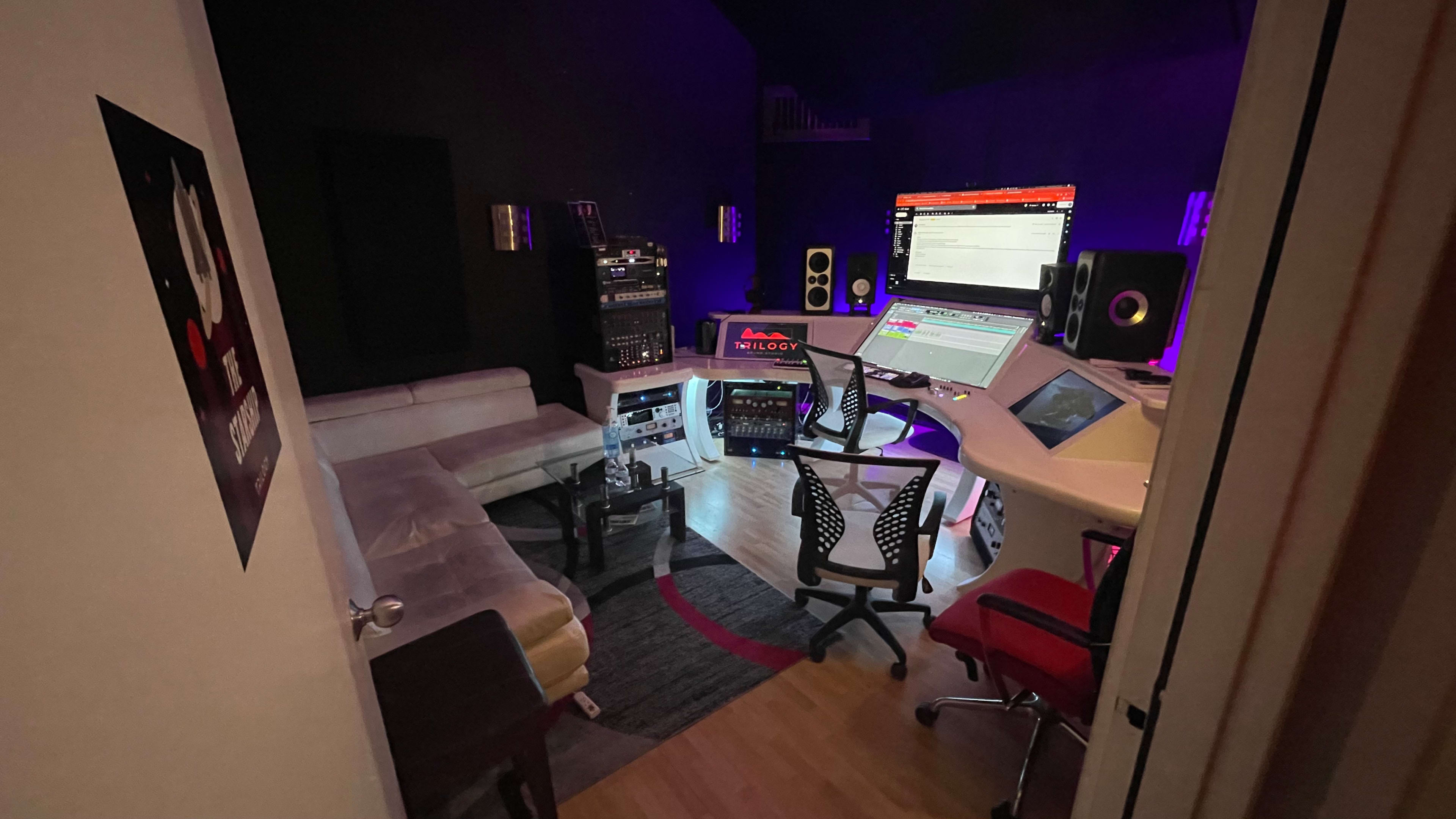 The image shows a dimly lit music studio with various audio equipment, two seating areas, and a large mixing console.