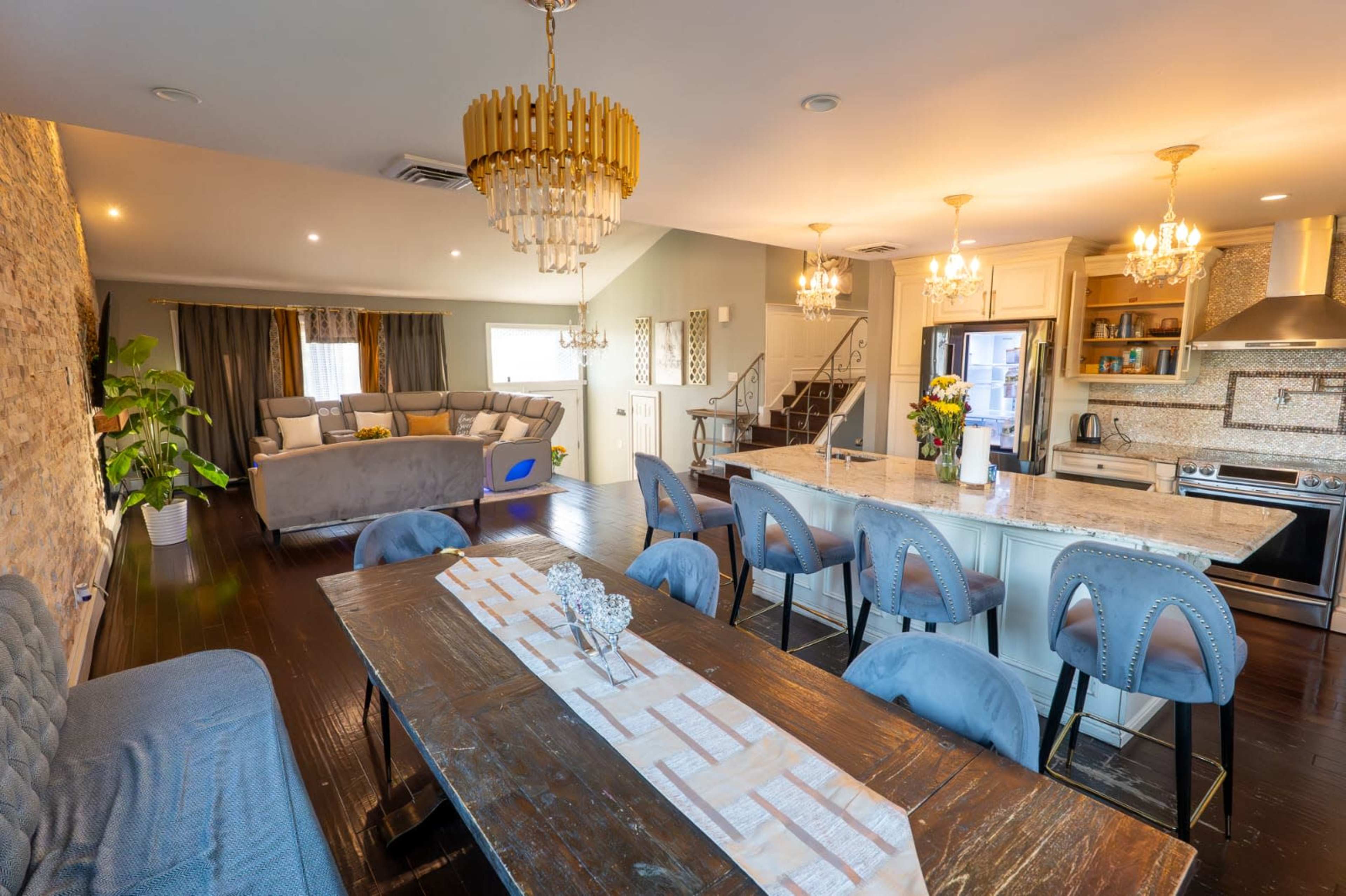 The image shows a modern open-concept living space featuring a dining area with a wooden table, a kitchen with an island, and a cozy seating area adorned with a chandelier and decorative lighting.