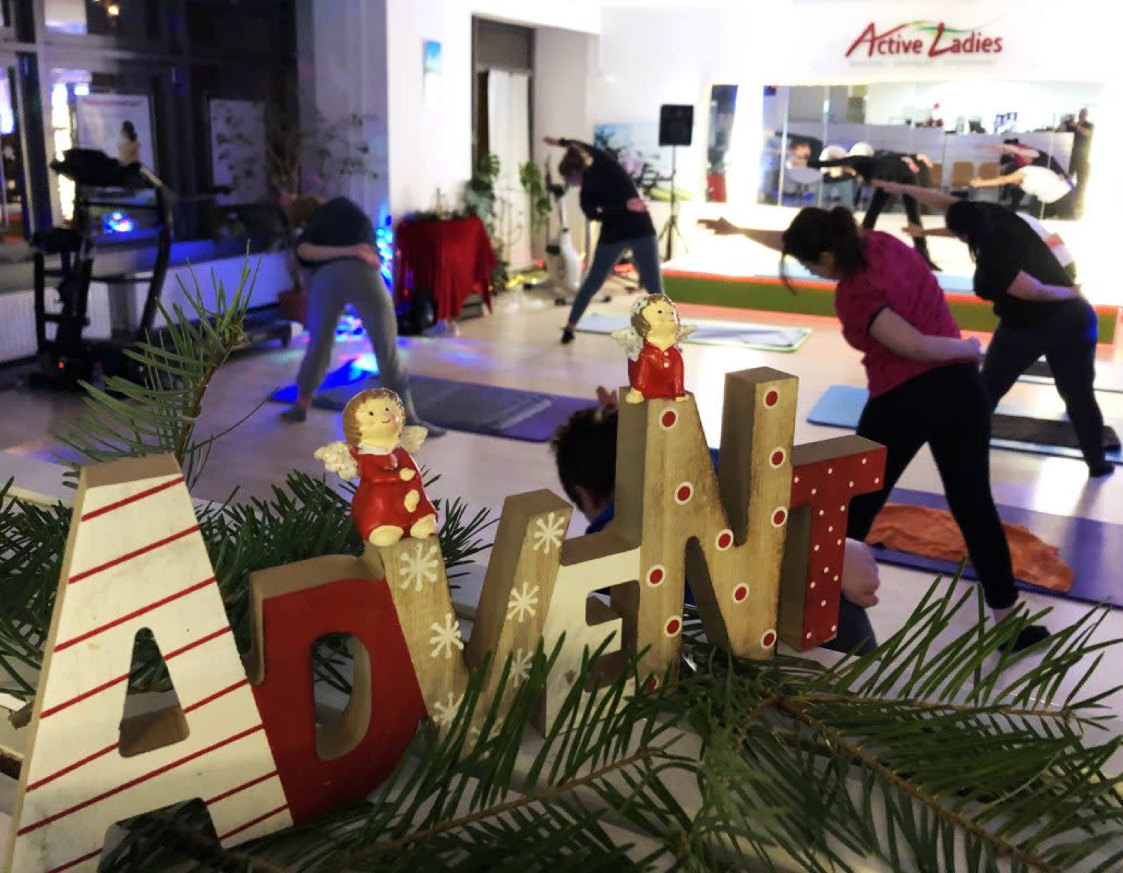 A decorated "ADVENT" sign is in the foreground while a group of women participates in a workout class in the background.
