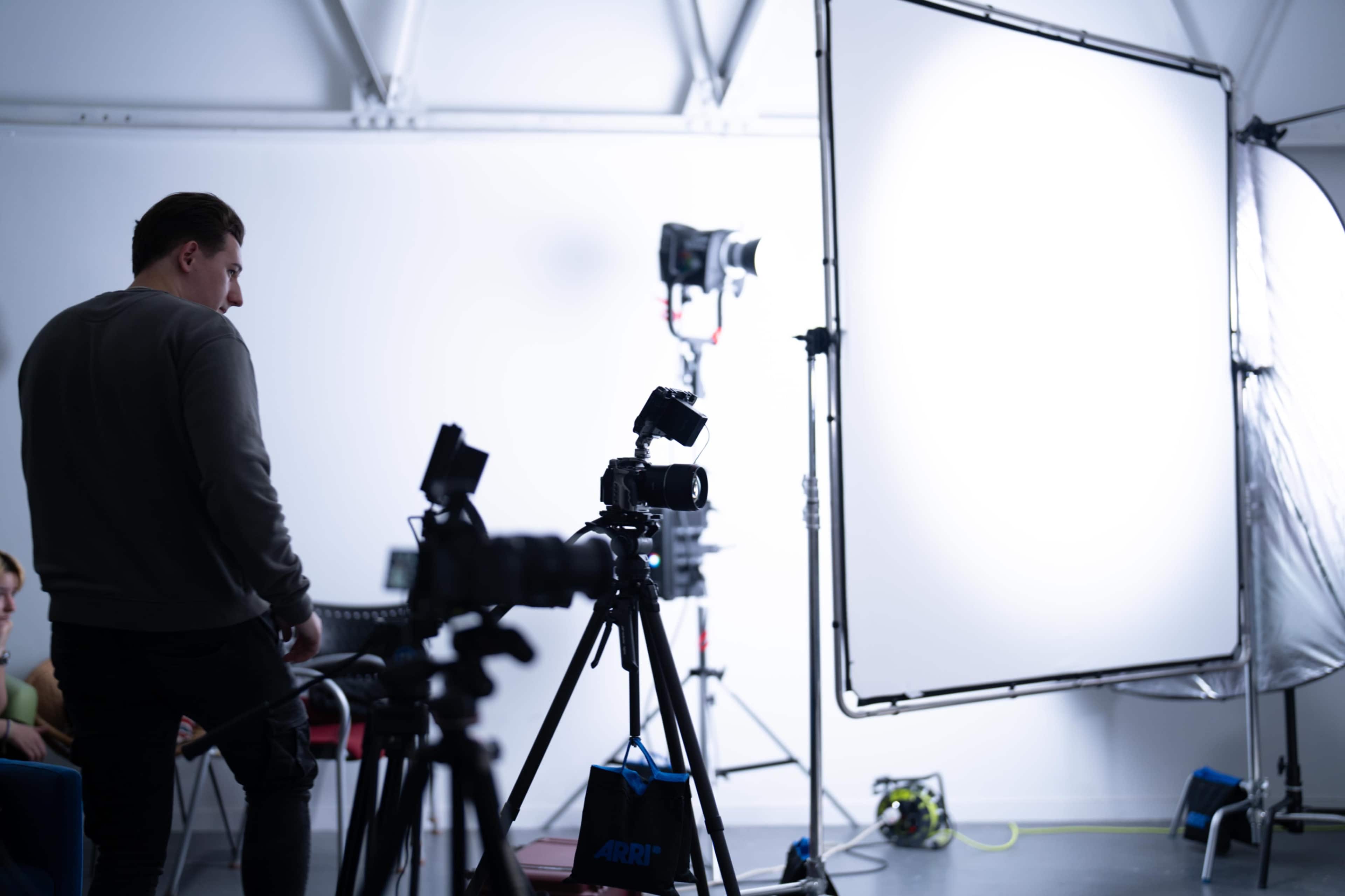 A man stands near a camera setup in a brightly lit studio, with a large white backdrop and additional lighting equipment visible.
