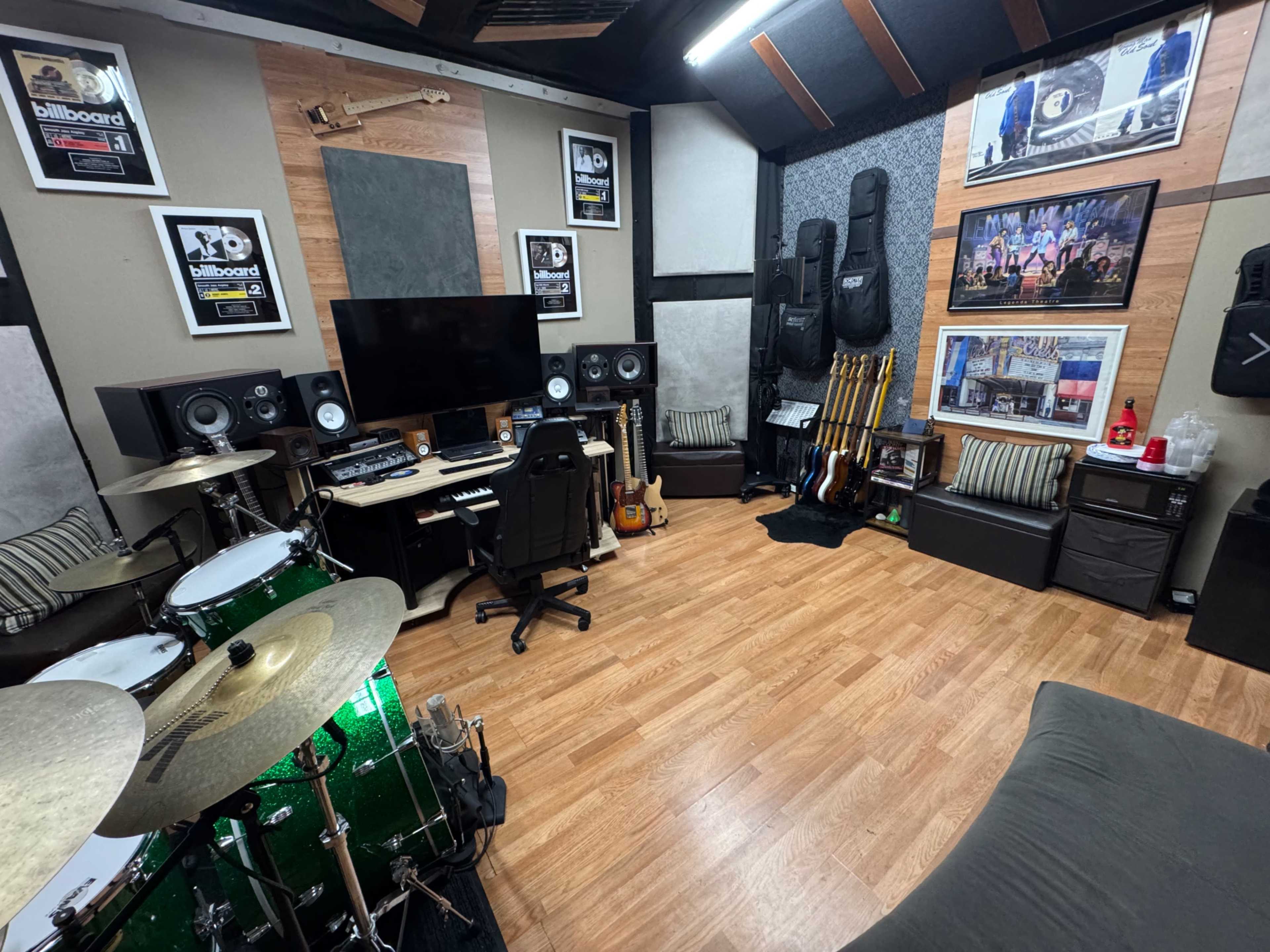 The image shows a music studio featuring a drum set, a computer workstation with speakers, various guitars displayed, and framed awards on the walls.