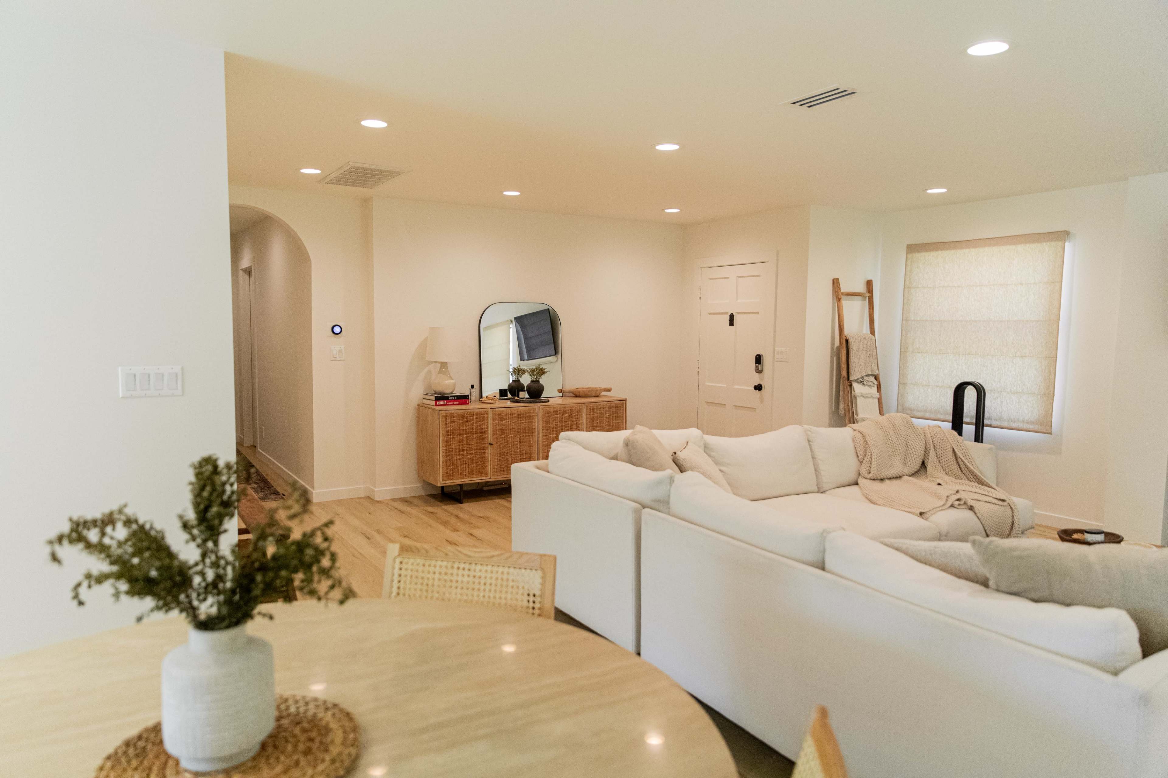 The image shows a modern living room with a white sectional sofa, light wood flooring, and natural light filtering through a window.