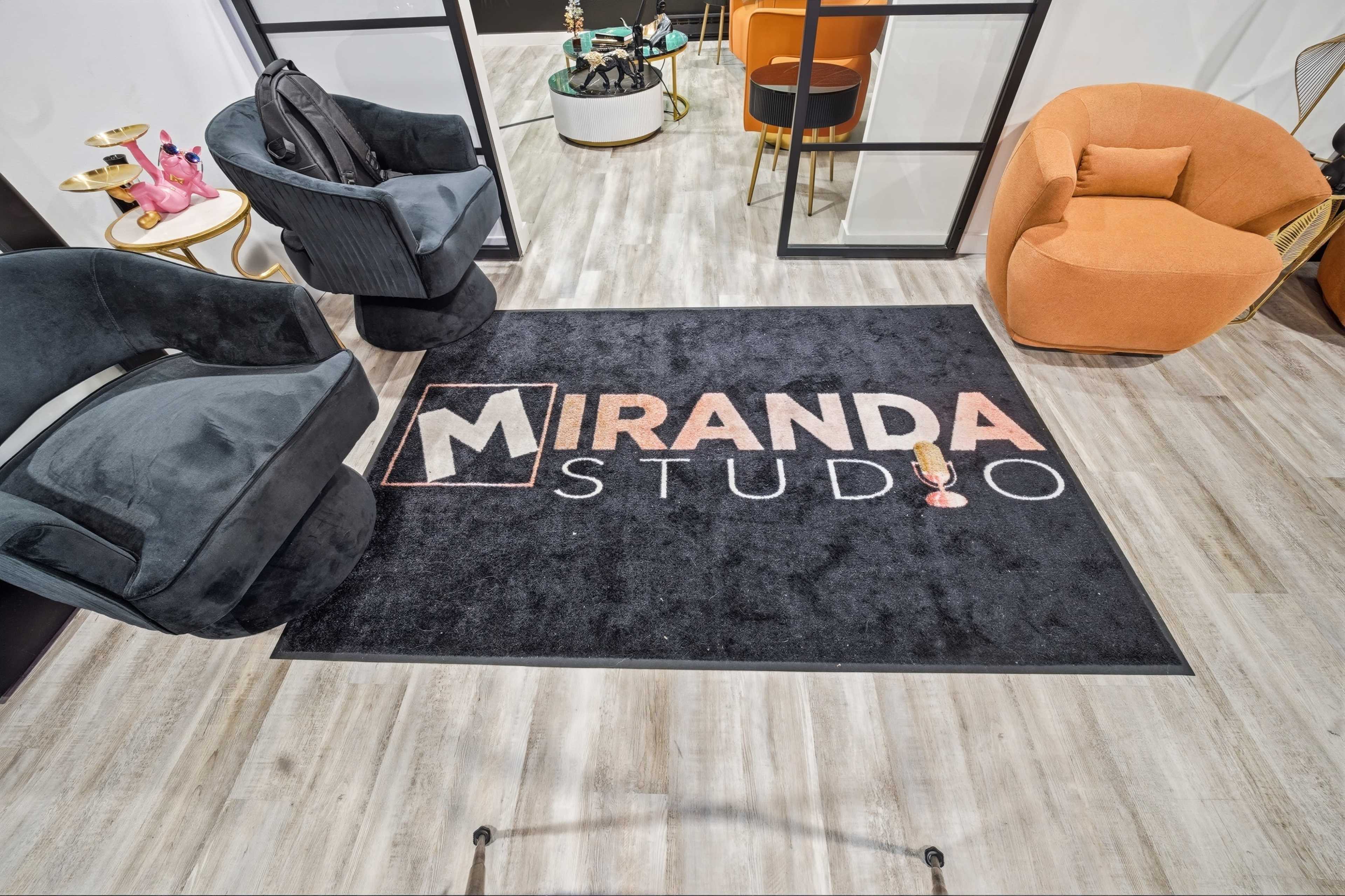 The image shows a stylish interior featuring a prominent black rug with the logo "MIRANDA STUDIO" in the center, surrounded by modern furniture including two armchairs.