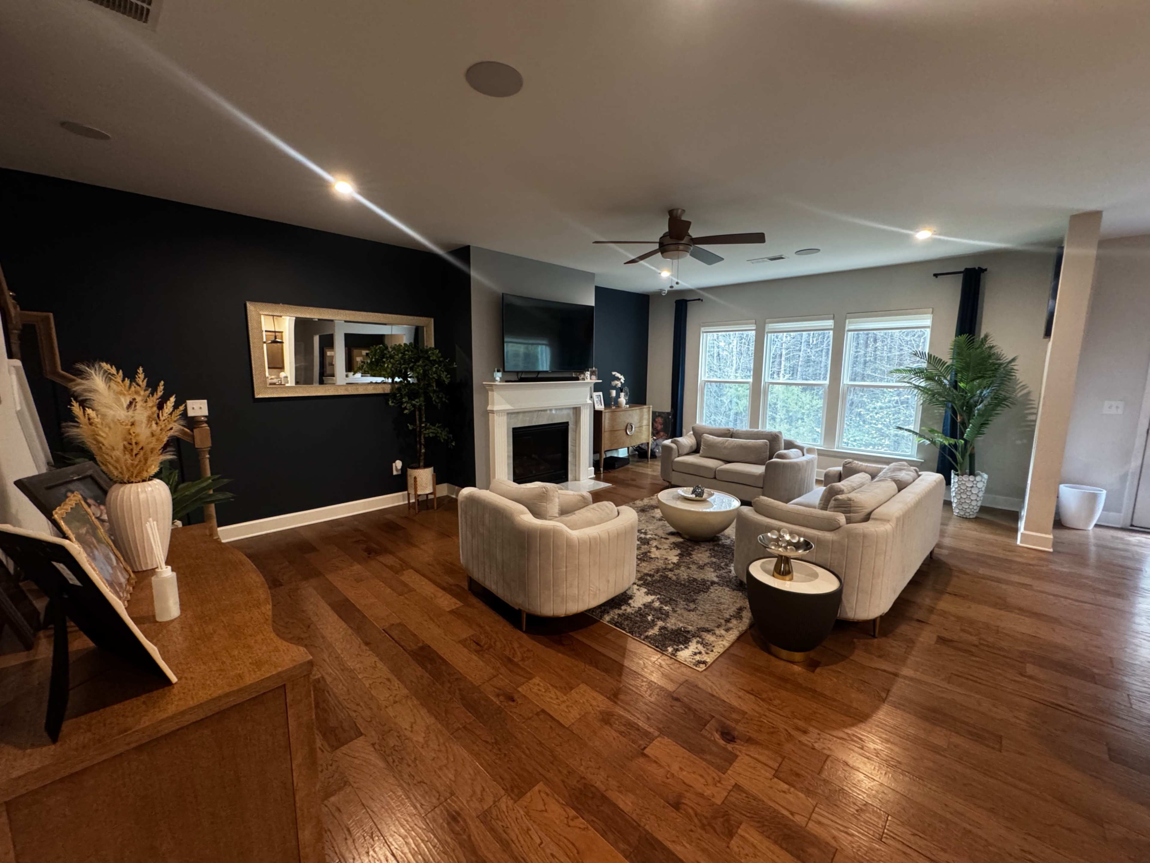 The living room features a centered seating arrangement with light-colored sofas, a flat-screen TV above a fireplace, and large windows overlooking a wooded area.