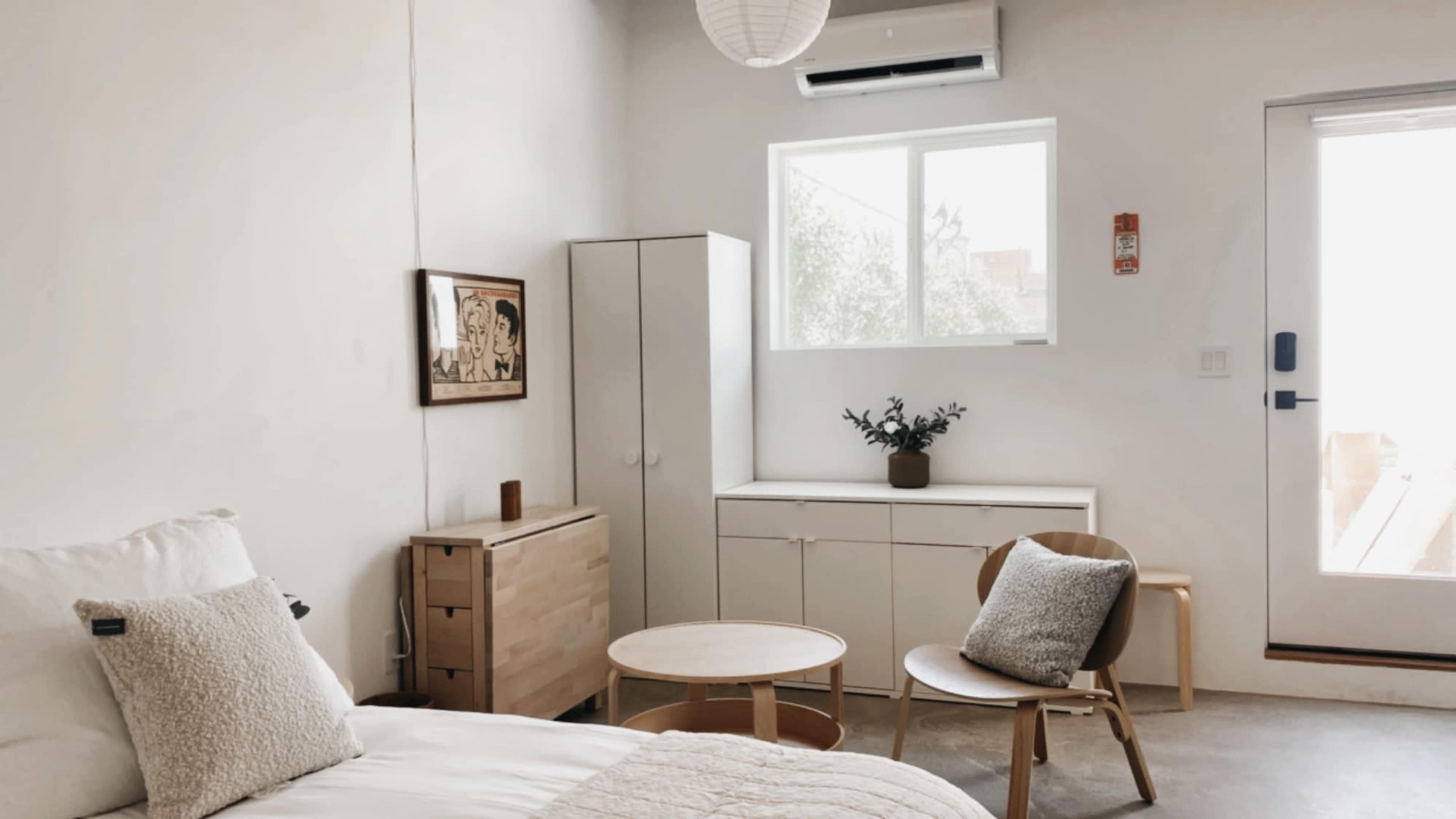 A minimalist bedroom features a bed, a wooden dresser, a cabinet, and a small table with a chair near a window and door.