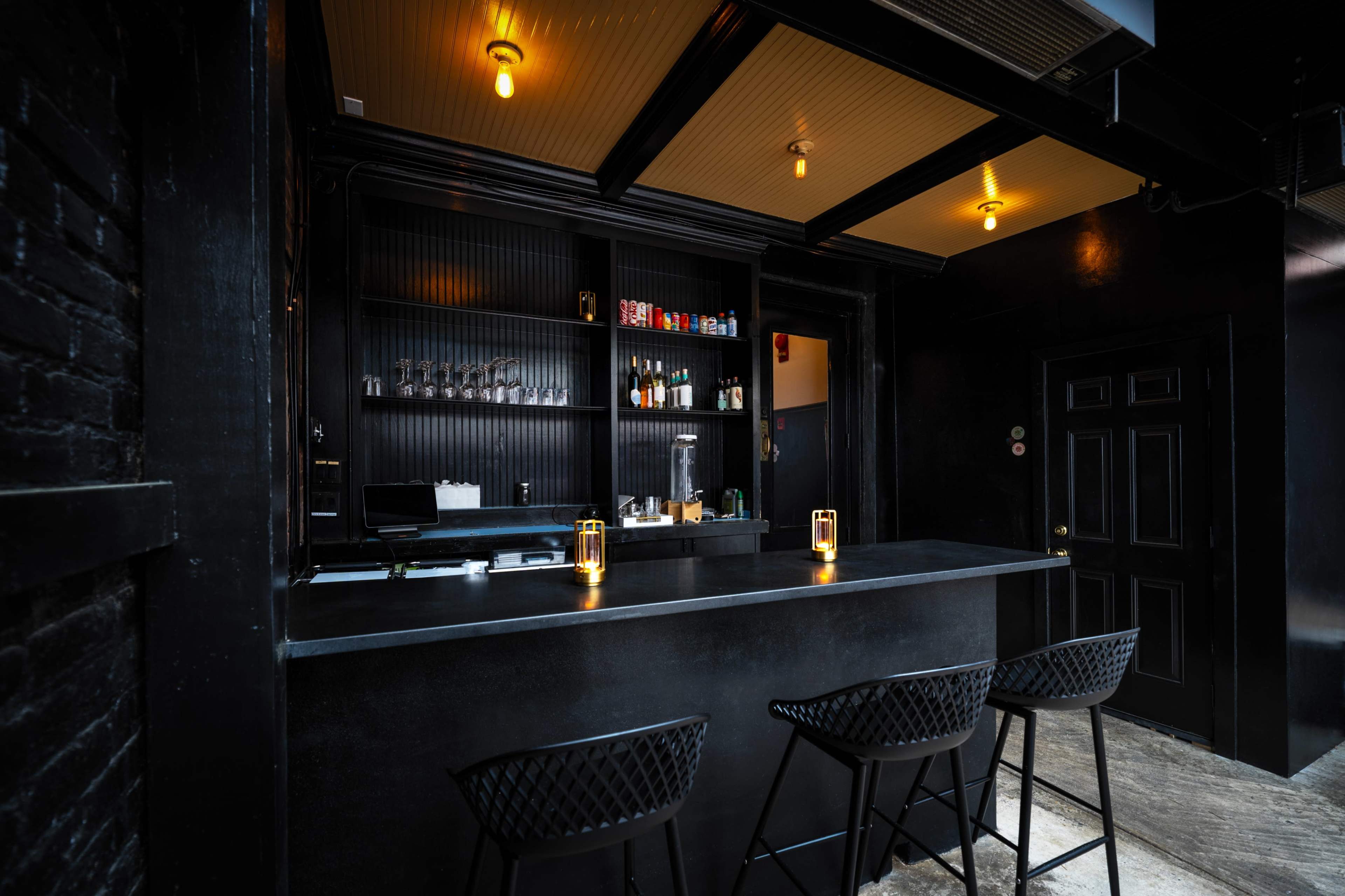 A dimly lit bar features a black counter and shelves stocked with various bottles, complemented by three high-backed stools.