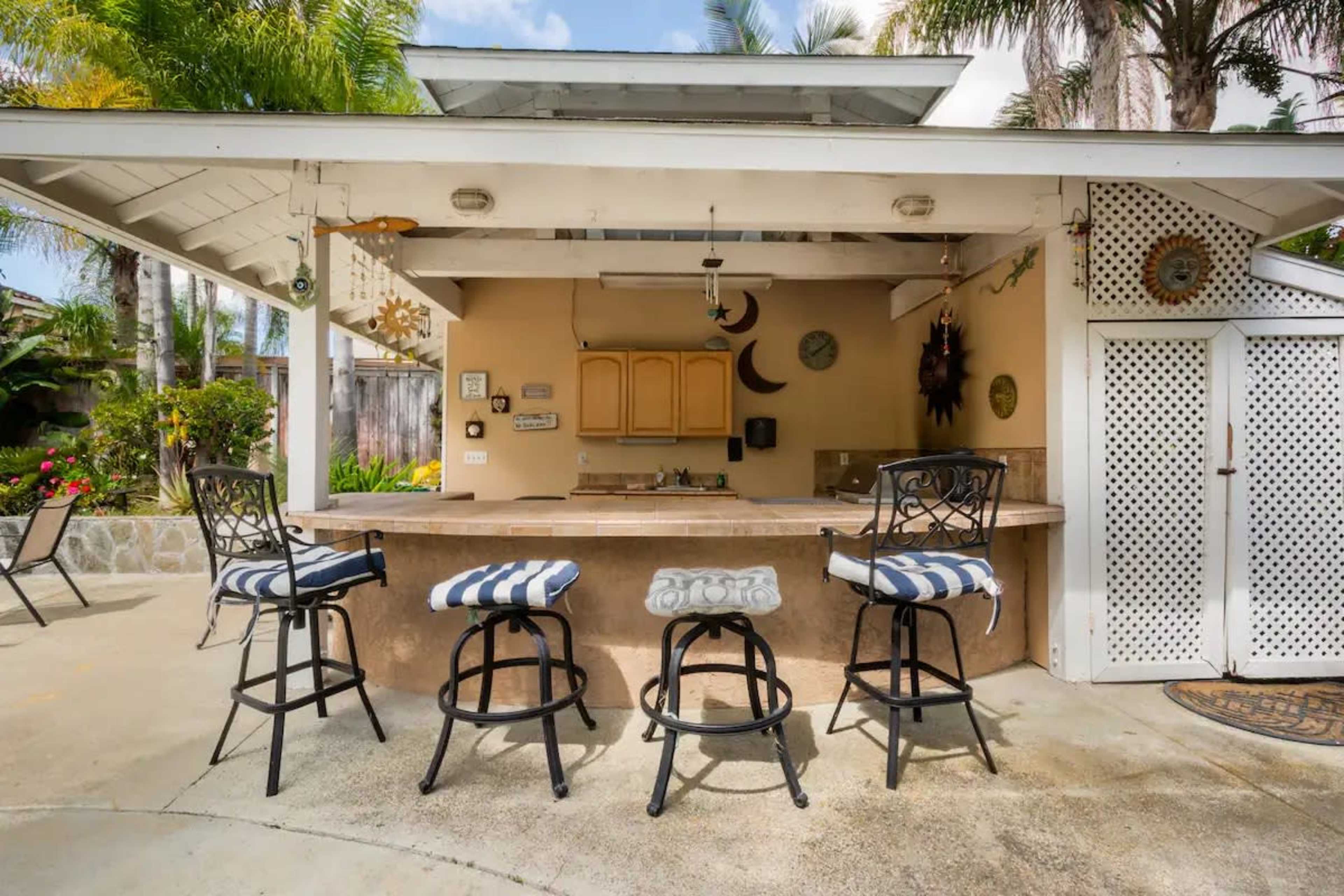 The image shows an outdoor bar area with four high stools, a countertop, and tropical greenery surrounding the space.