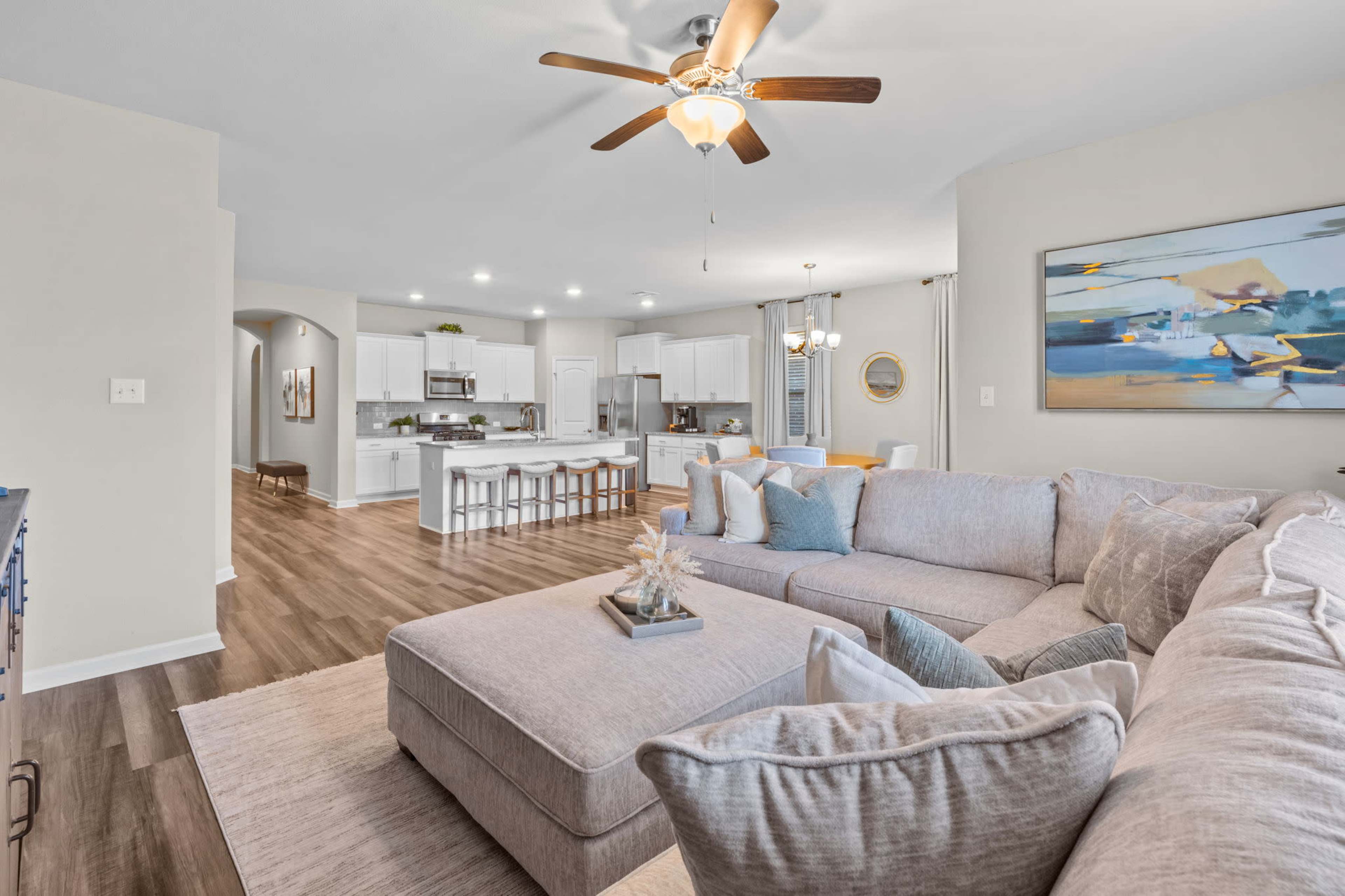 A spacious living area with a sectional sofa, ceiling fan, and an open kitchen visible in the background.
