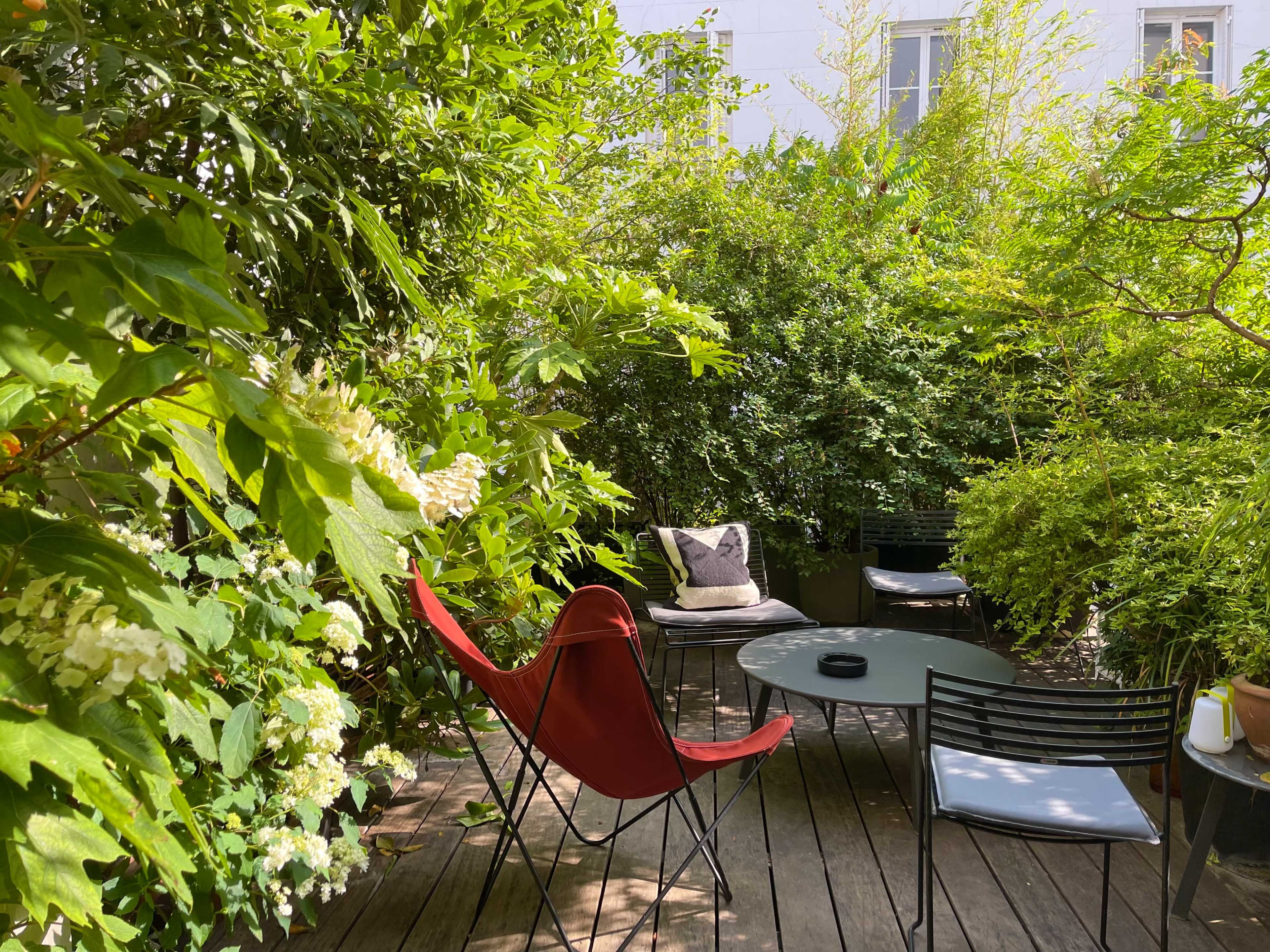 A wooden deck is surrounded by dense green foliage, featuring a red chair and a circular table with two additional black chairs.