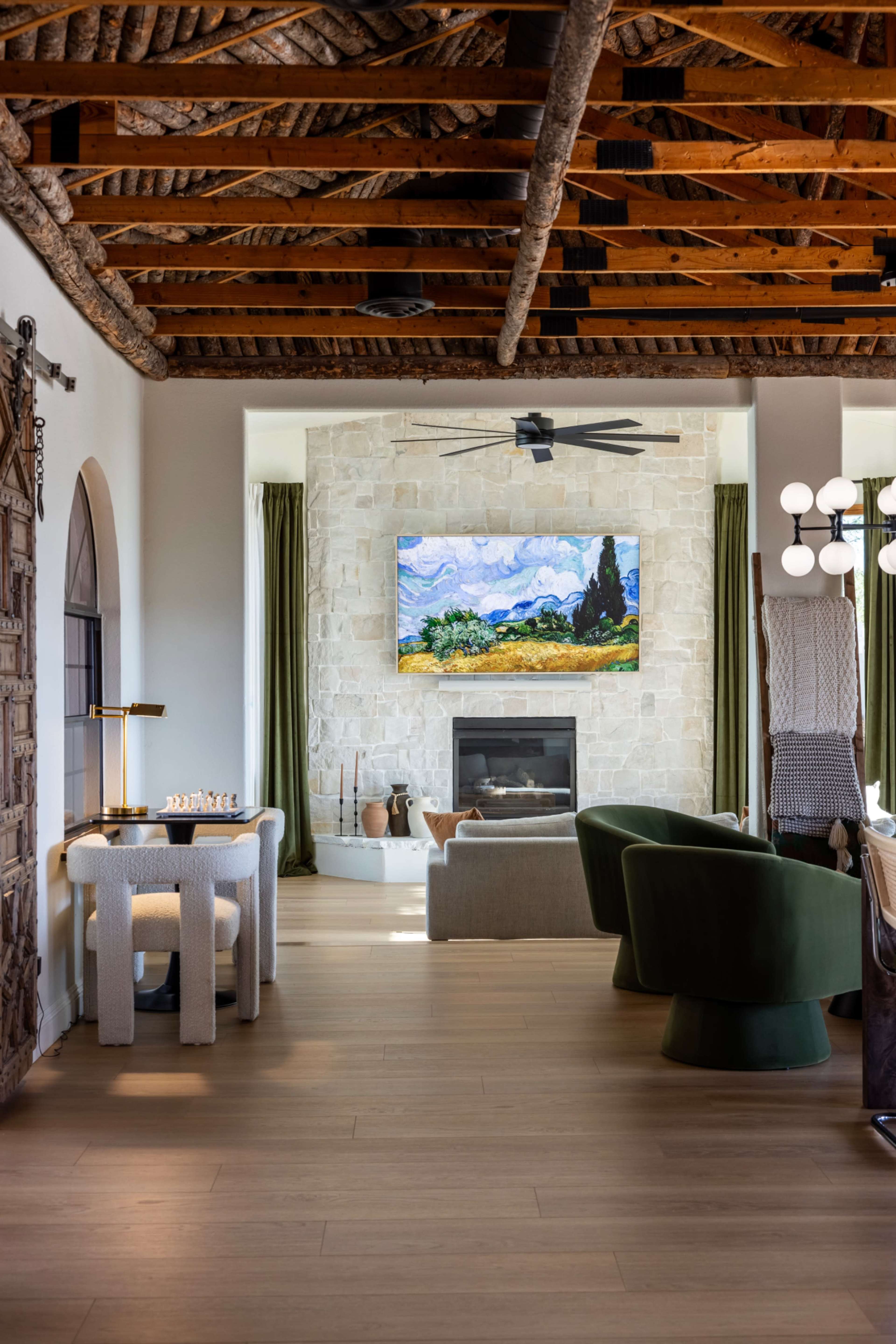 The image shows a modern living room featuring a stone fireplace, a mounted artwork, green accent chairs, and a wooden beam ceiling.