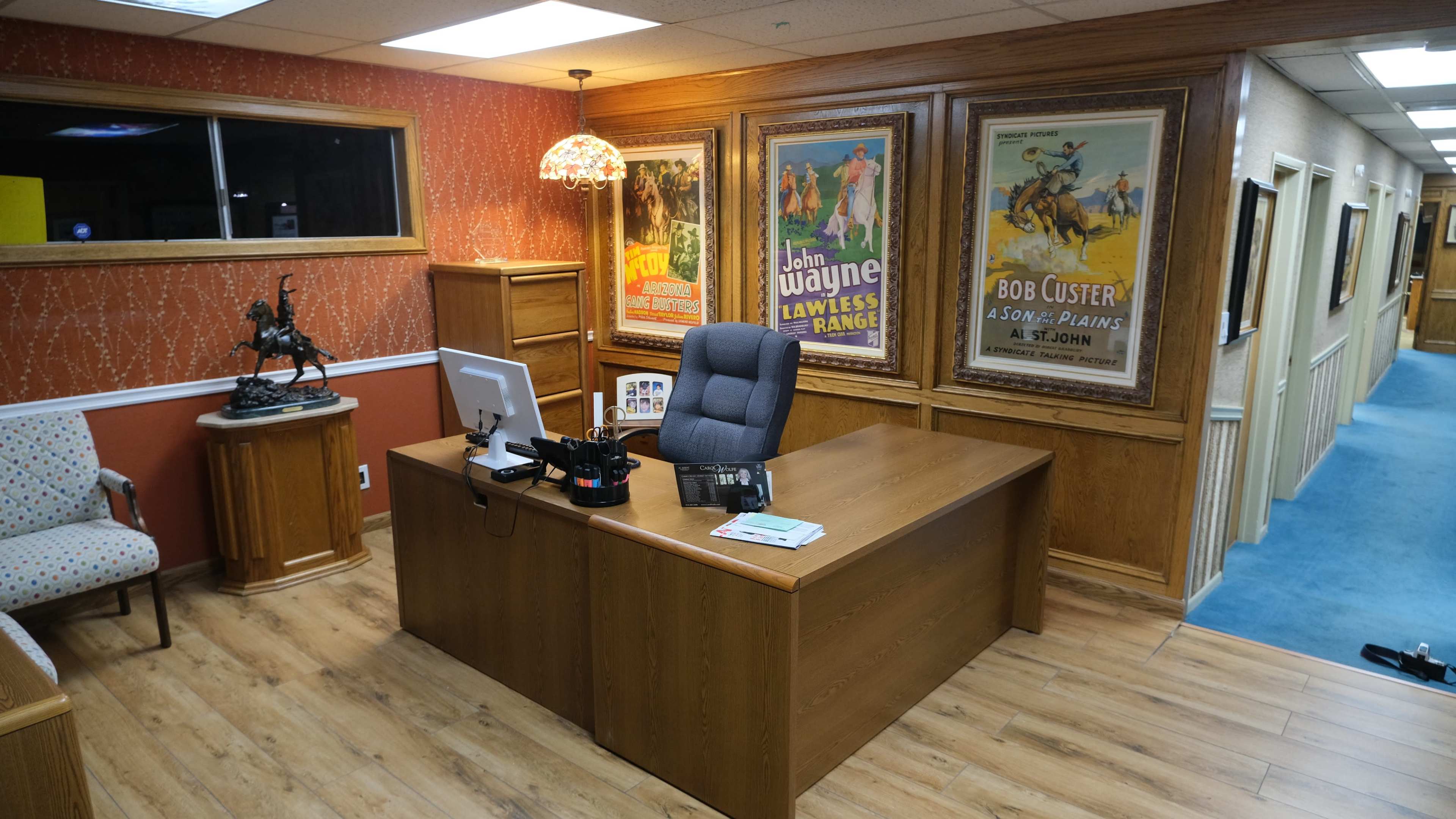 An office space with wooden furniture, a computer on a desk, vintage movie posters on the walls, and a small seating area with patterned upholstery.