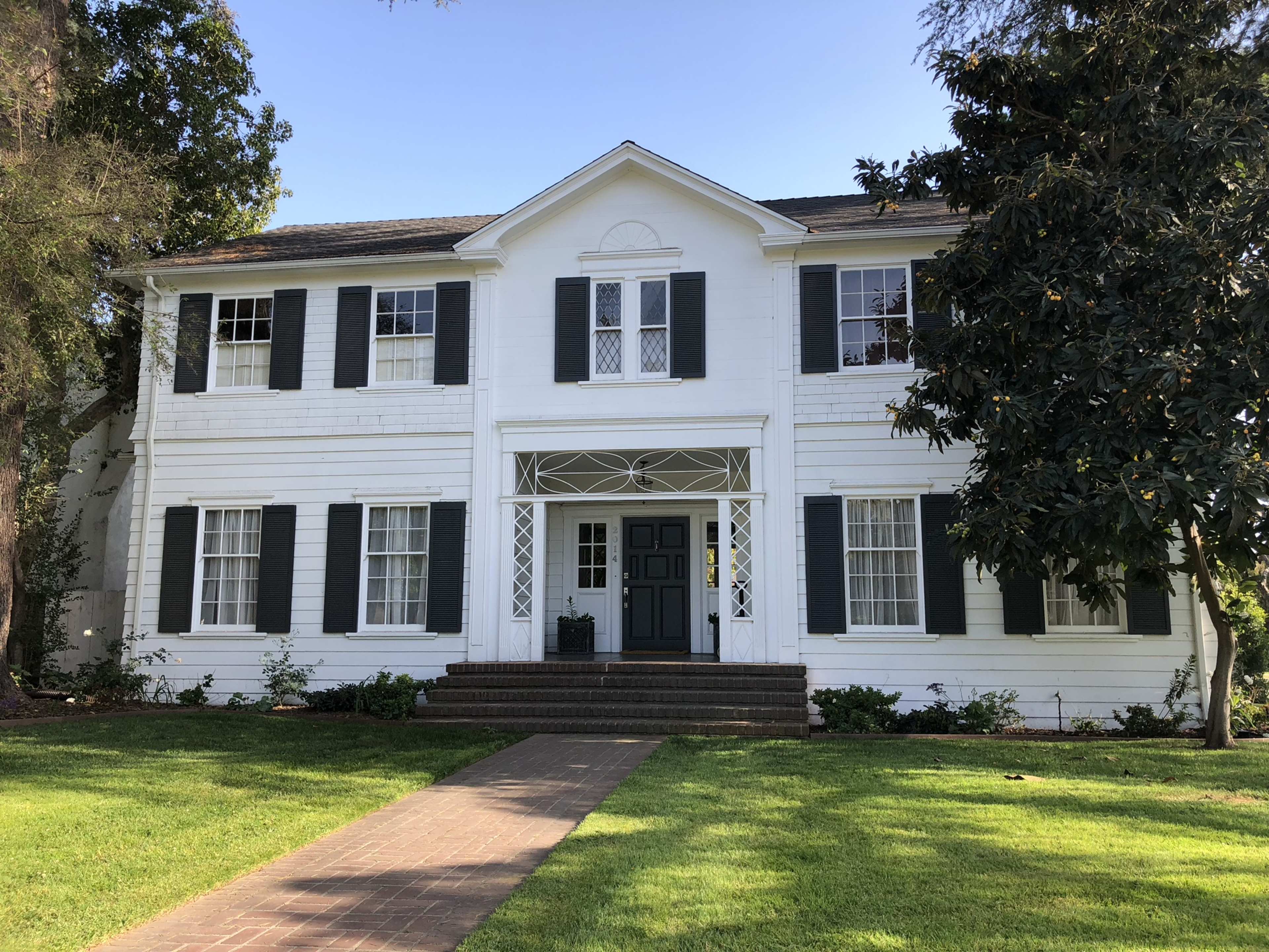 A two-story white house with green shutters and a front porch features a centered door and symmetrical window arrangement.