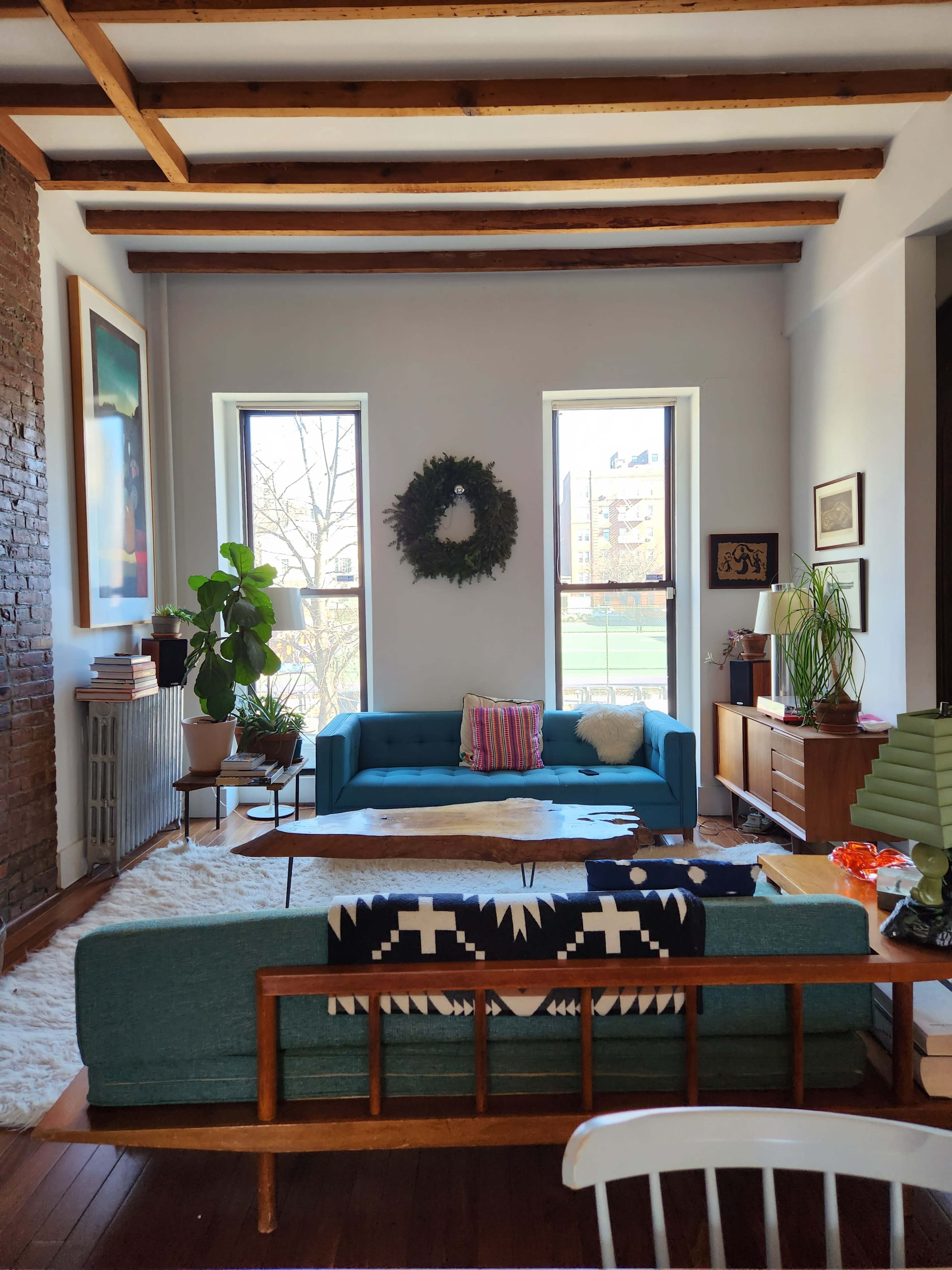 The living room features a blue sofa, wooden beams overhead, and two large windows surrounded by decorative elements.