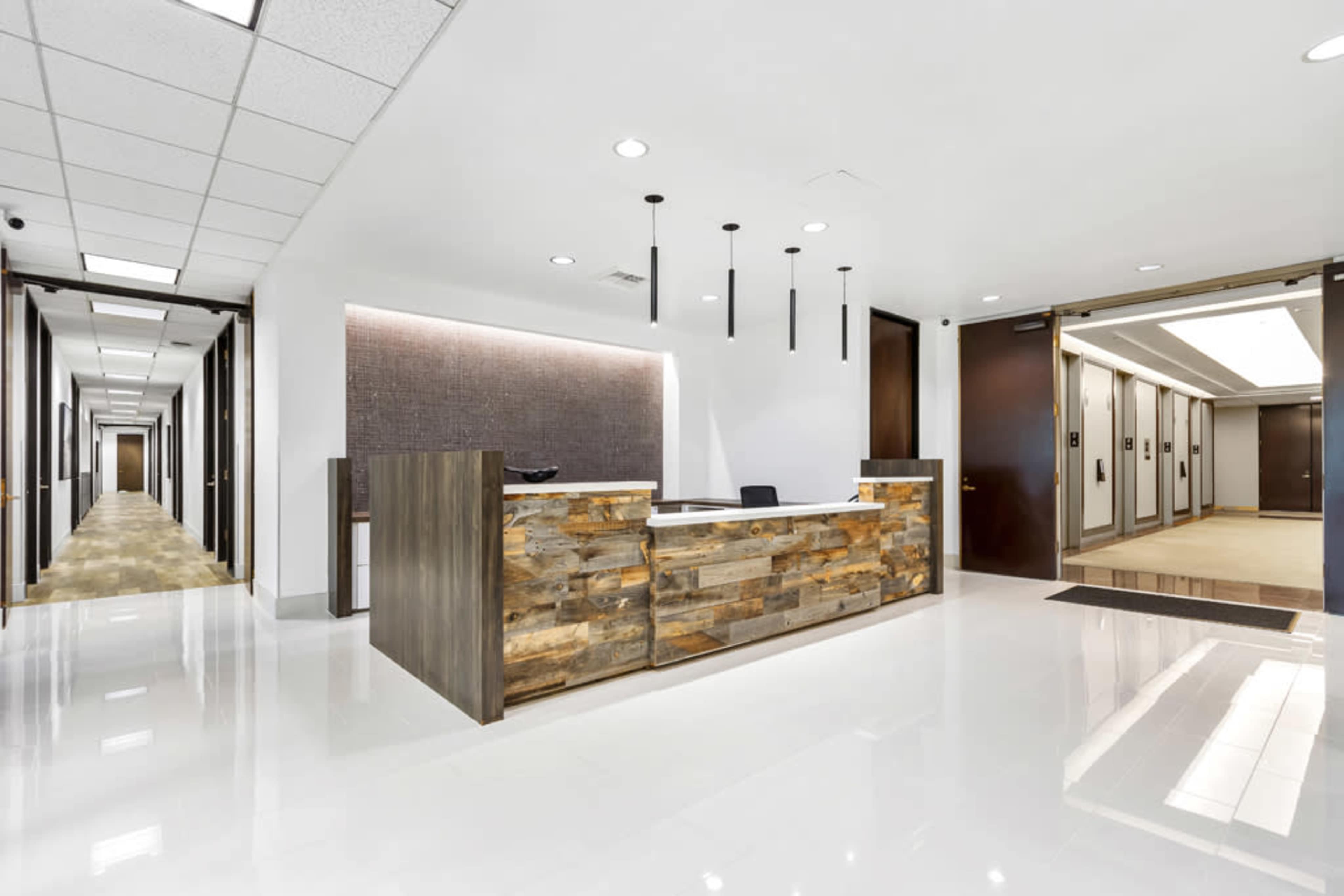 The image shows a modern reception area with a wooden front desk and a hallway leading to elevators and office doors.