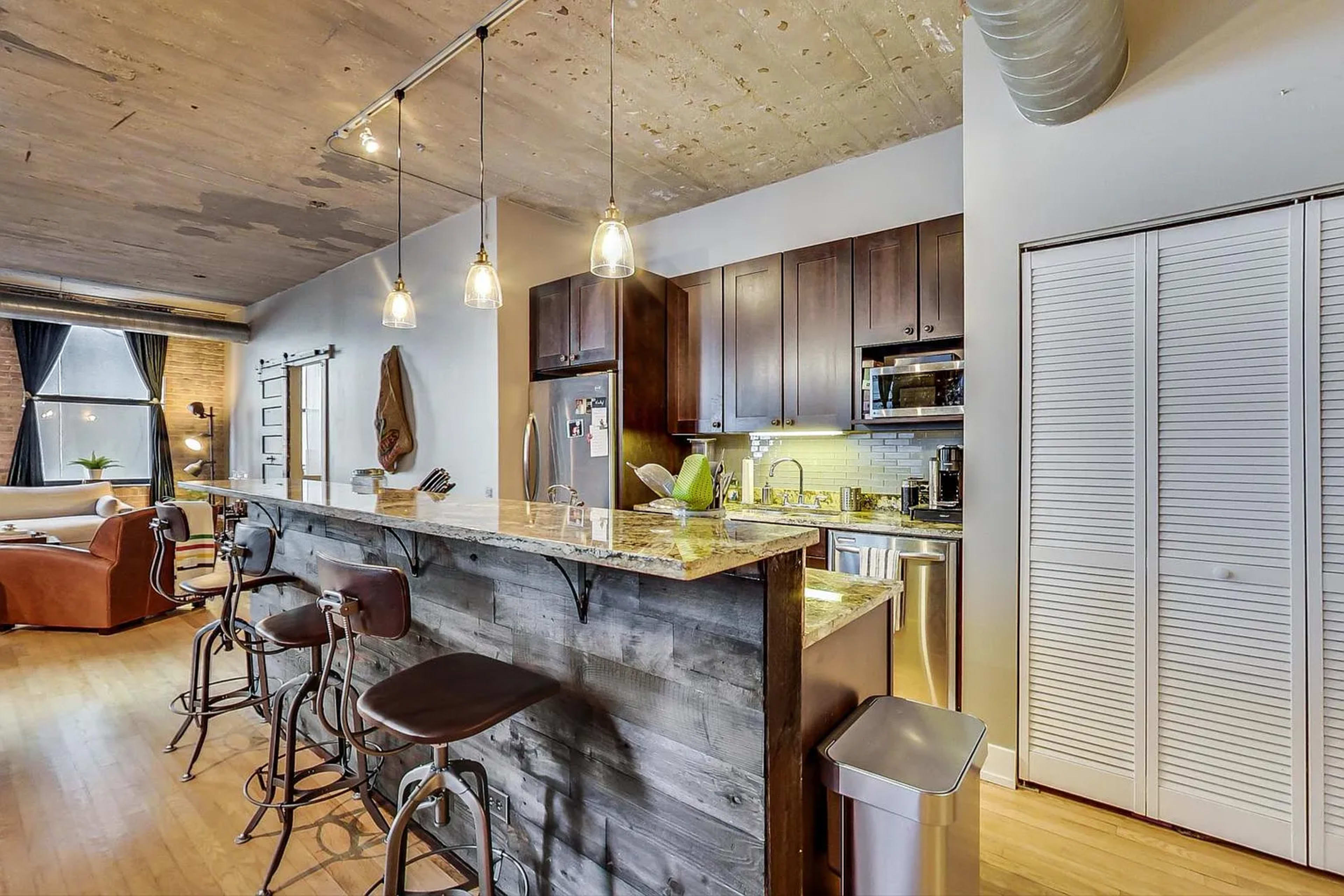 The image shows a kitchen with a wooden bar, three stools, hanging pendant lights, and modern appliances in an open-concept living space.