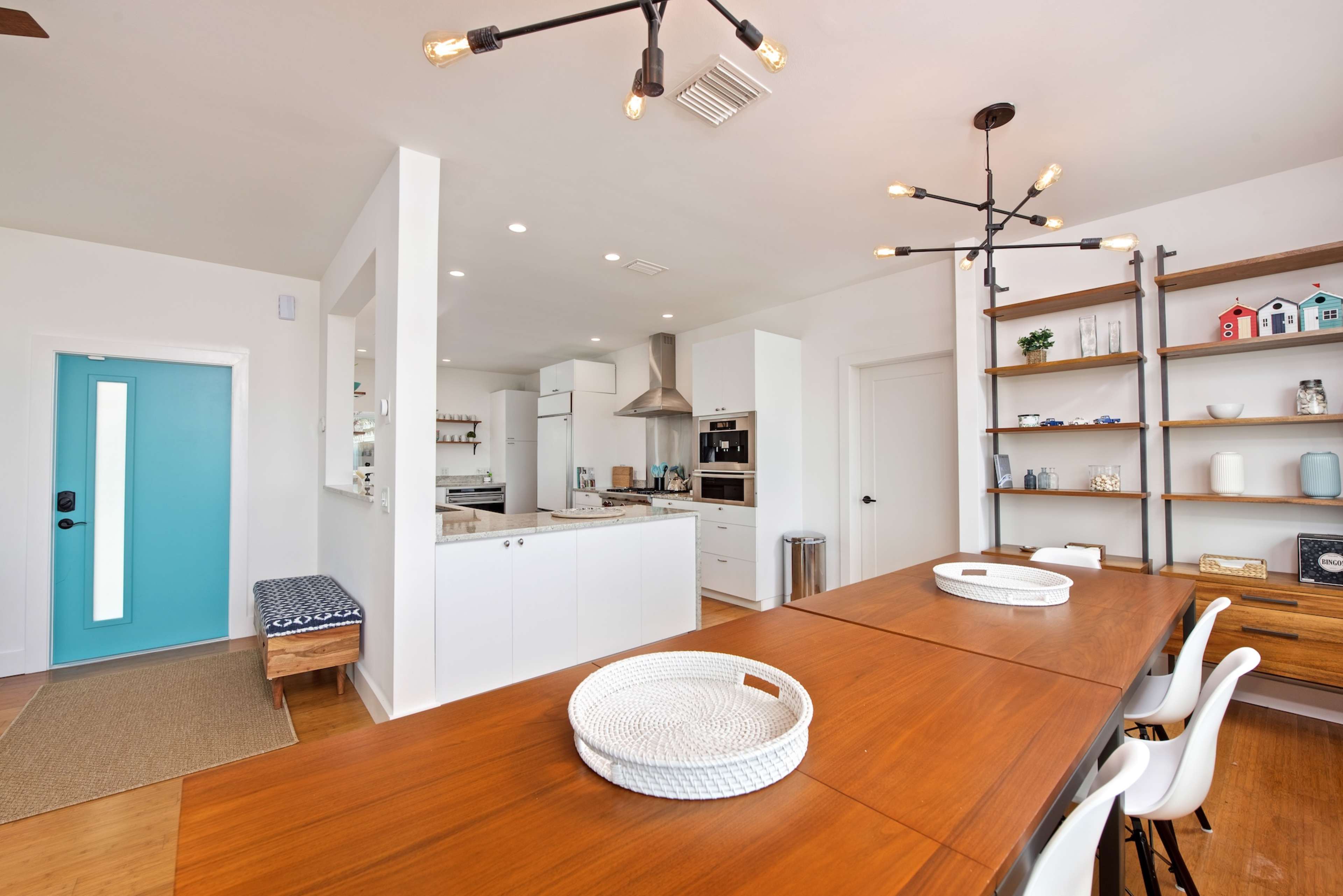 A modern kitchen and dining area features a long wooden table, white cabinetry, and a turquoise door leading outside.