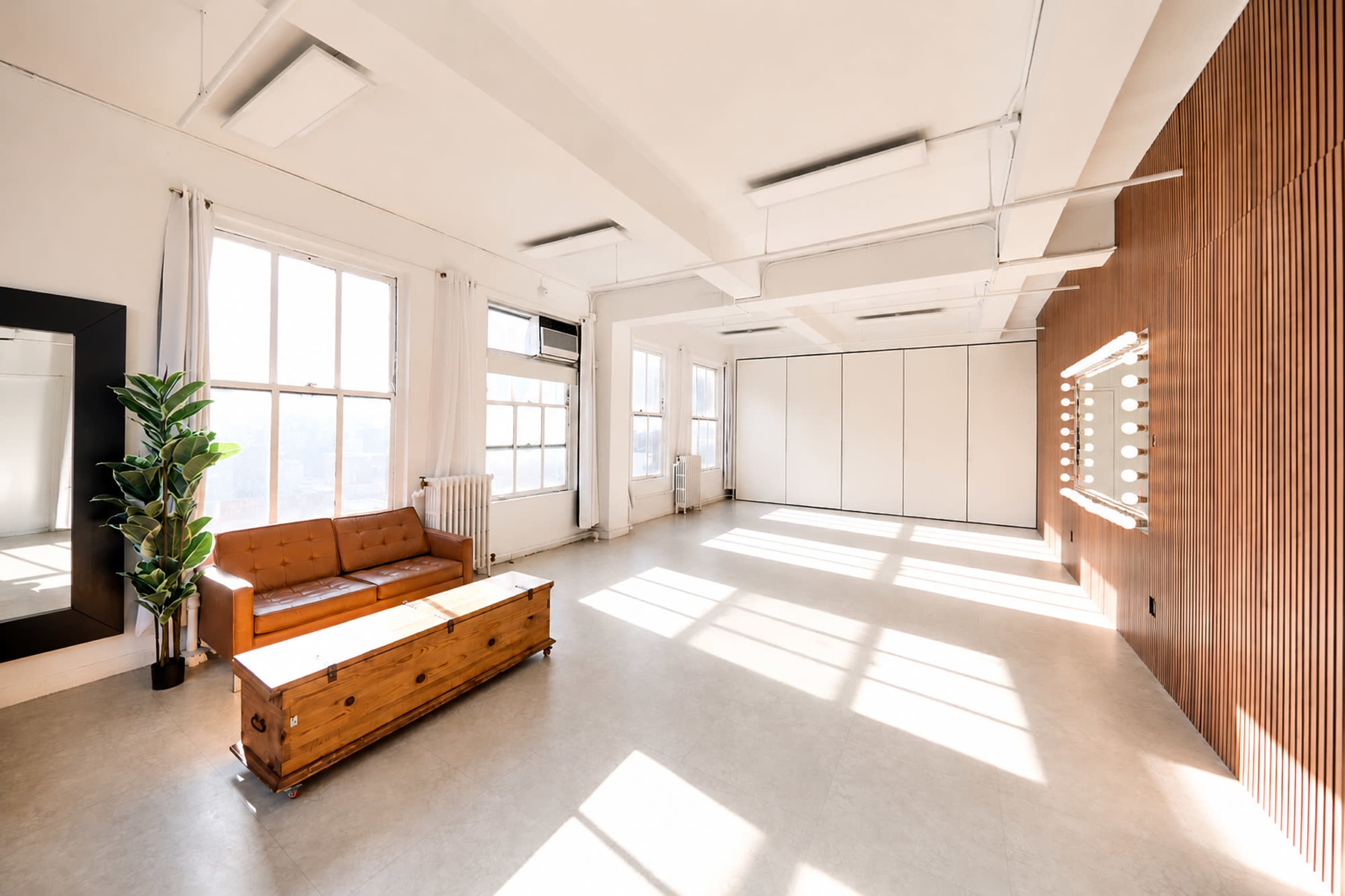 The image shows a spacious room with large windows, a wooden couch, a vintage wooden trunk, and a wall featuring a row of bright lights.