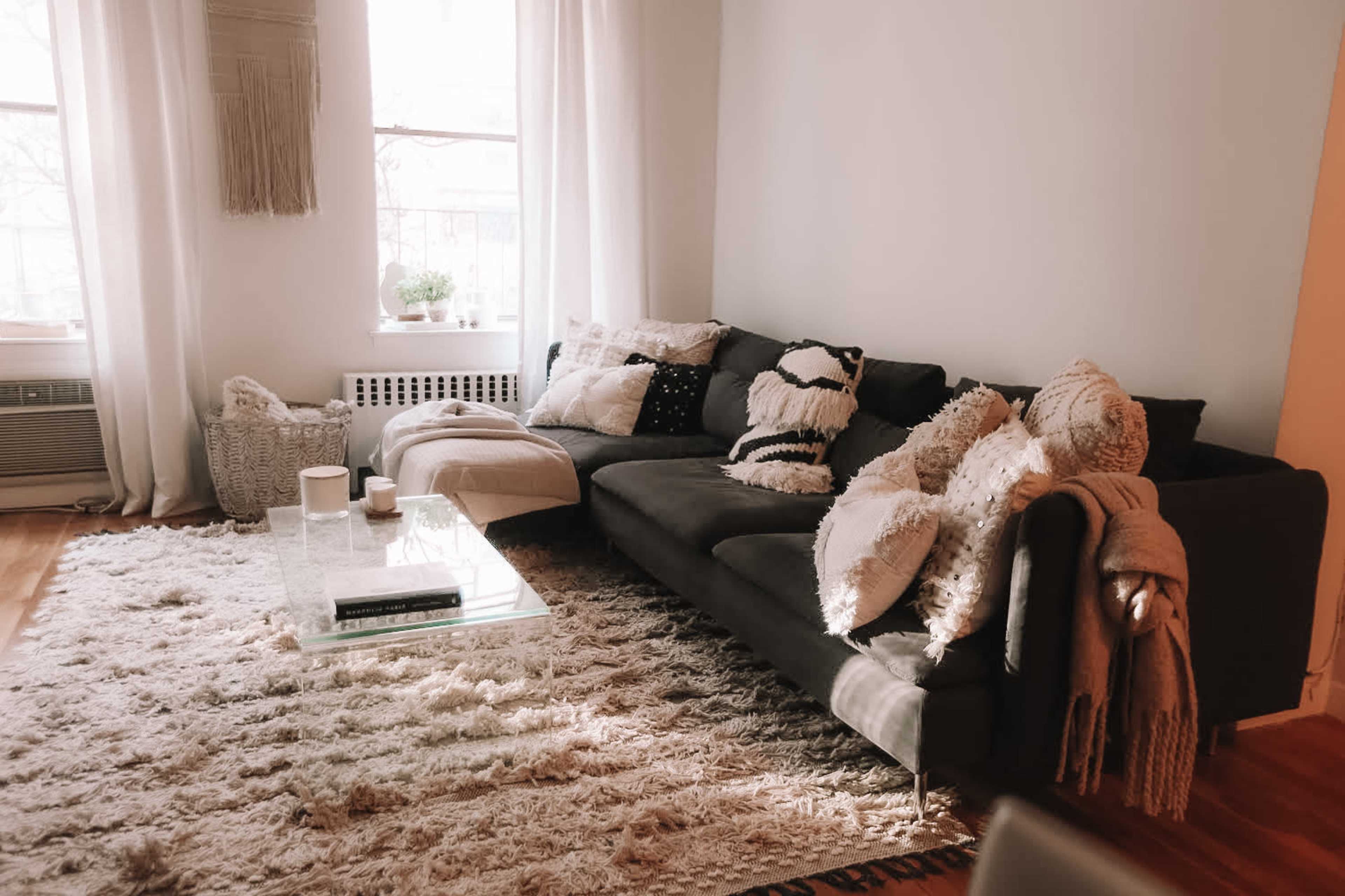 A dark sectional sofa with various decorative pillows sits on a plush rug in a well-lit living room with large windows.