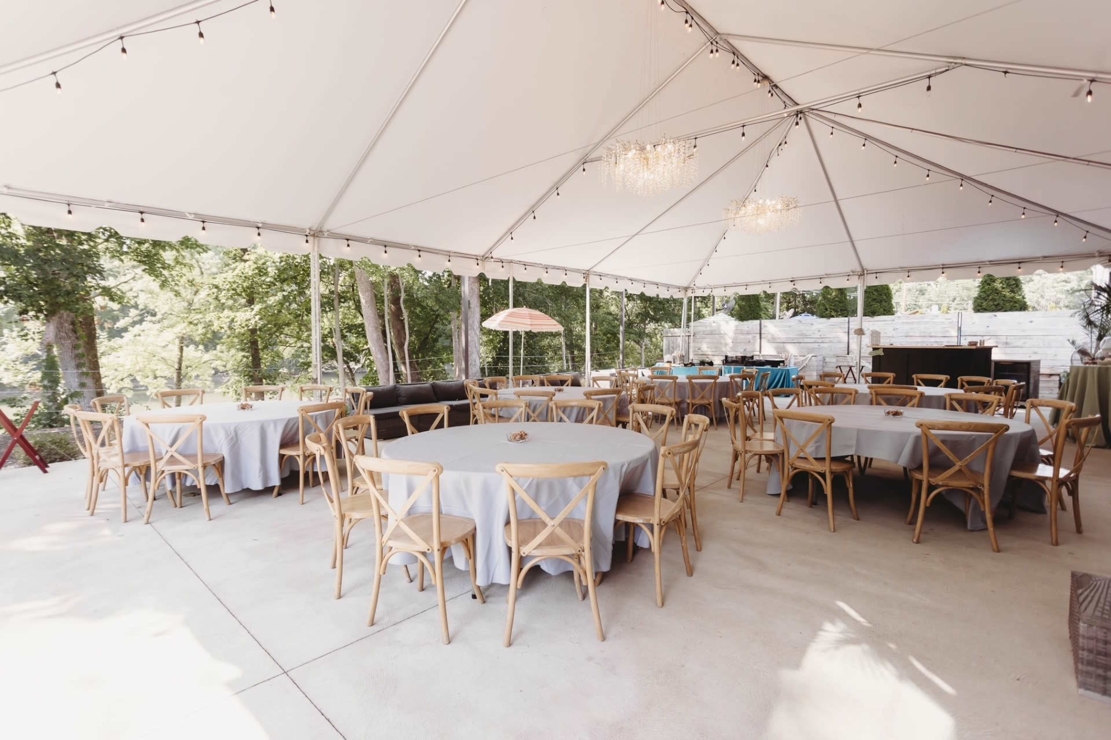 A large outdoor tent is set up with multiple round tables and wooden chairs, surrounded by greenery.