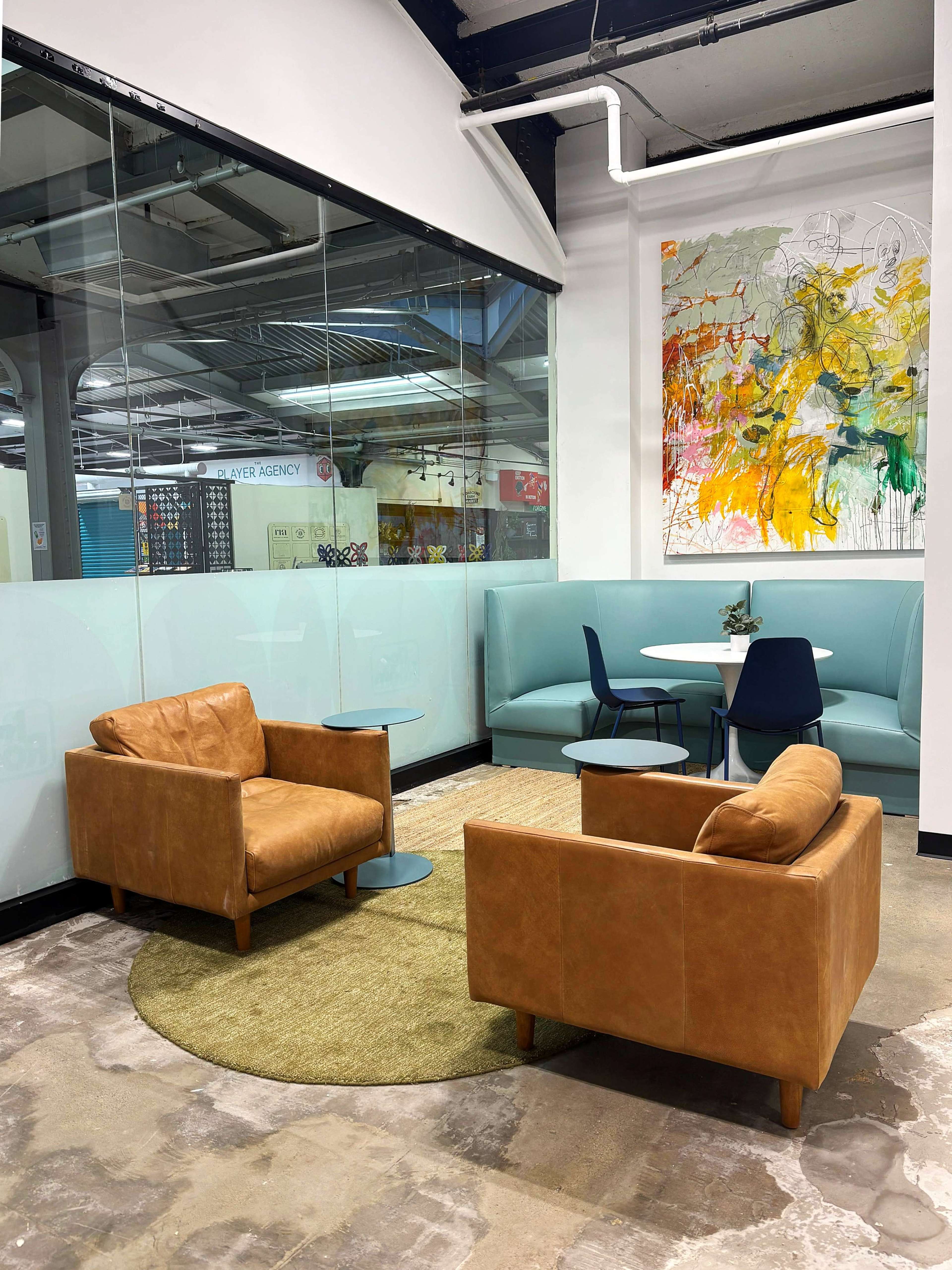 The image shows a modern seating area with two brown leather chairs on a round rug, a small table, and a colorful abstract artwork on the wall.