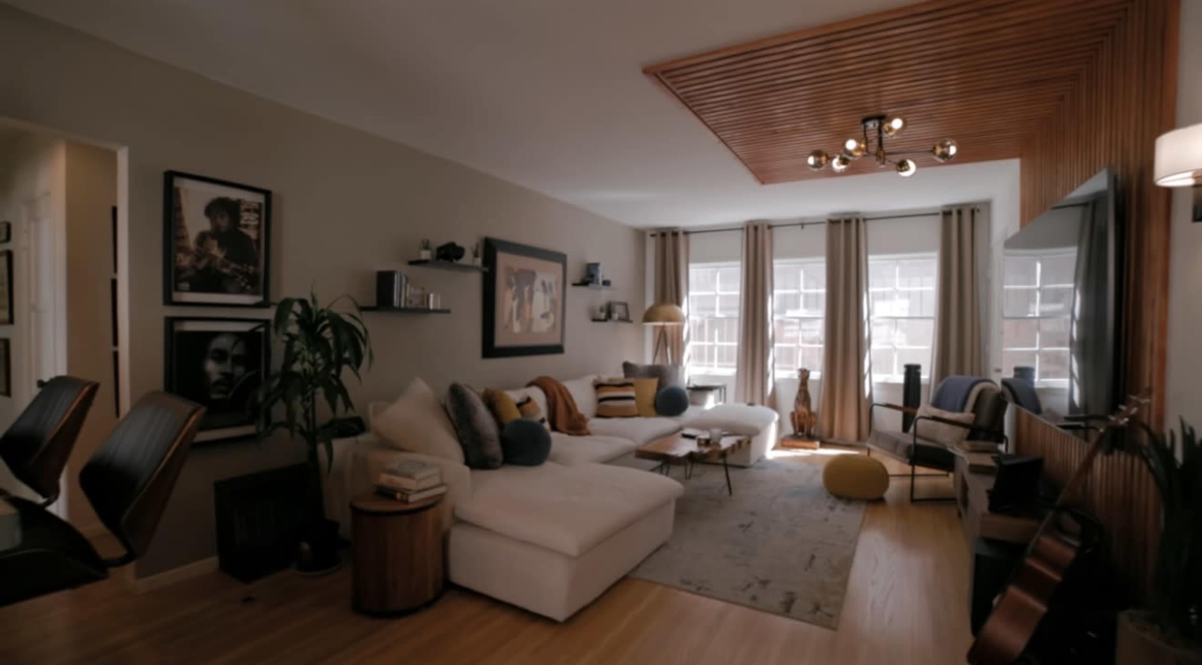 The image shows a modern living room with a white sectional sofa, a wooden ceiling feature, and large windows dressed with light curtains.