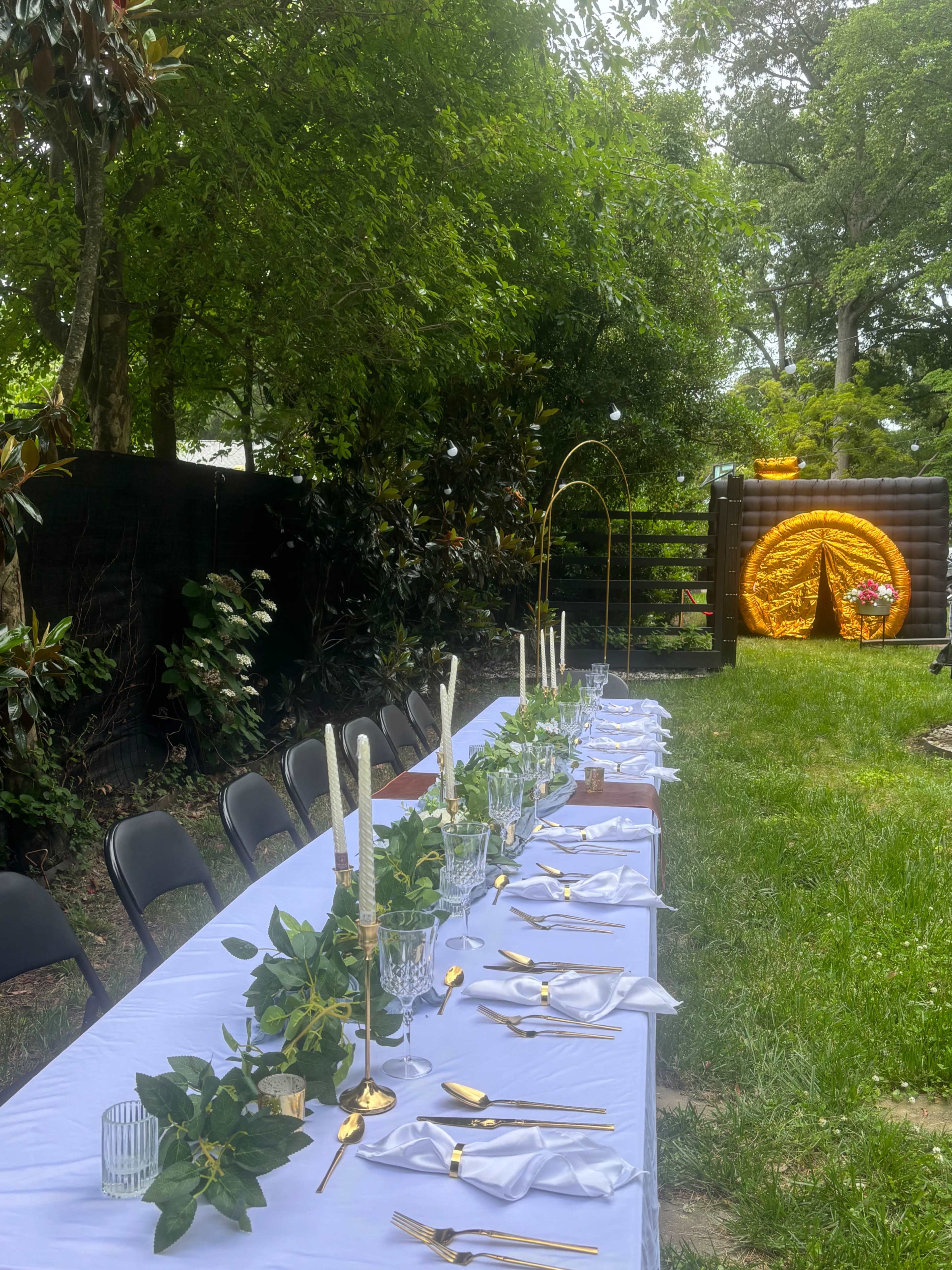 A long dining table is set outdoors with white linens, gold utensils, and greenery, positioned beside a decorative circular backdrop in a garden setting.