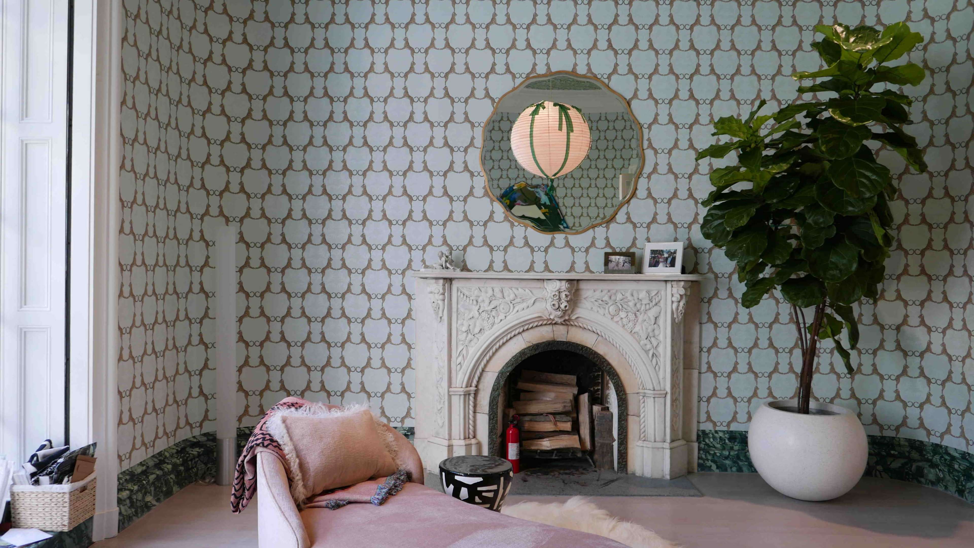 The image features a room with patterned wallpaper, a round mirror above a white fireplace, a plant in a pot, and a small table beside a cozy seating area.