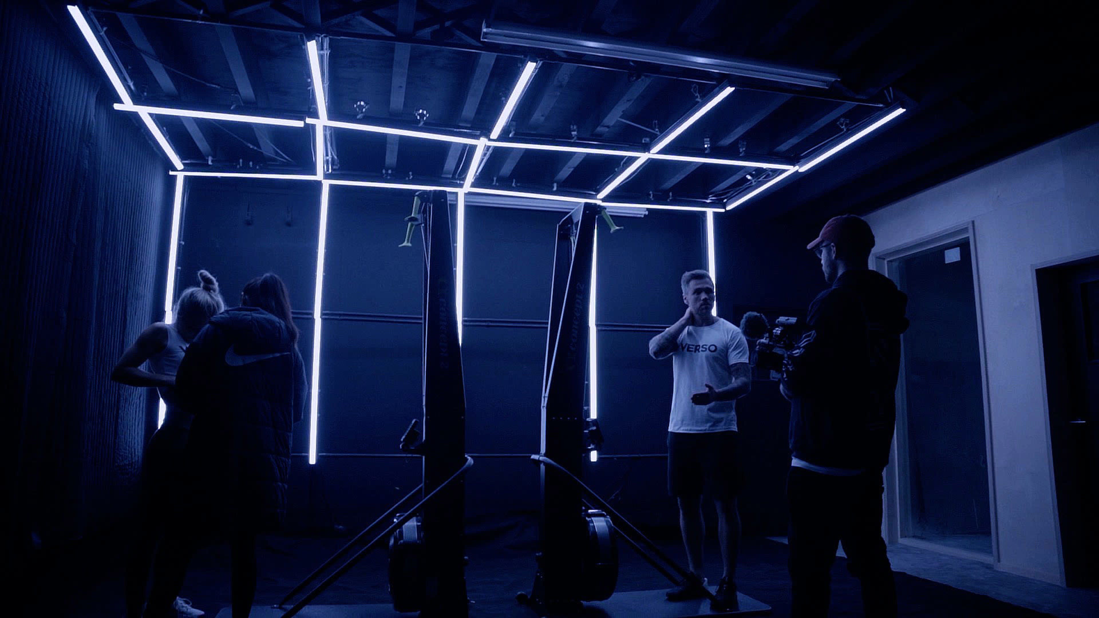 A dark, minimalist gym space with two rowing machines and individuals interacting under illuminated geometric lines.