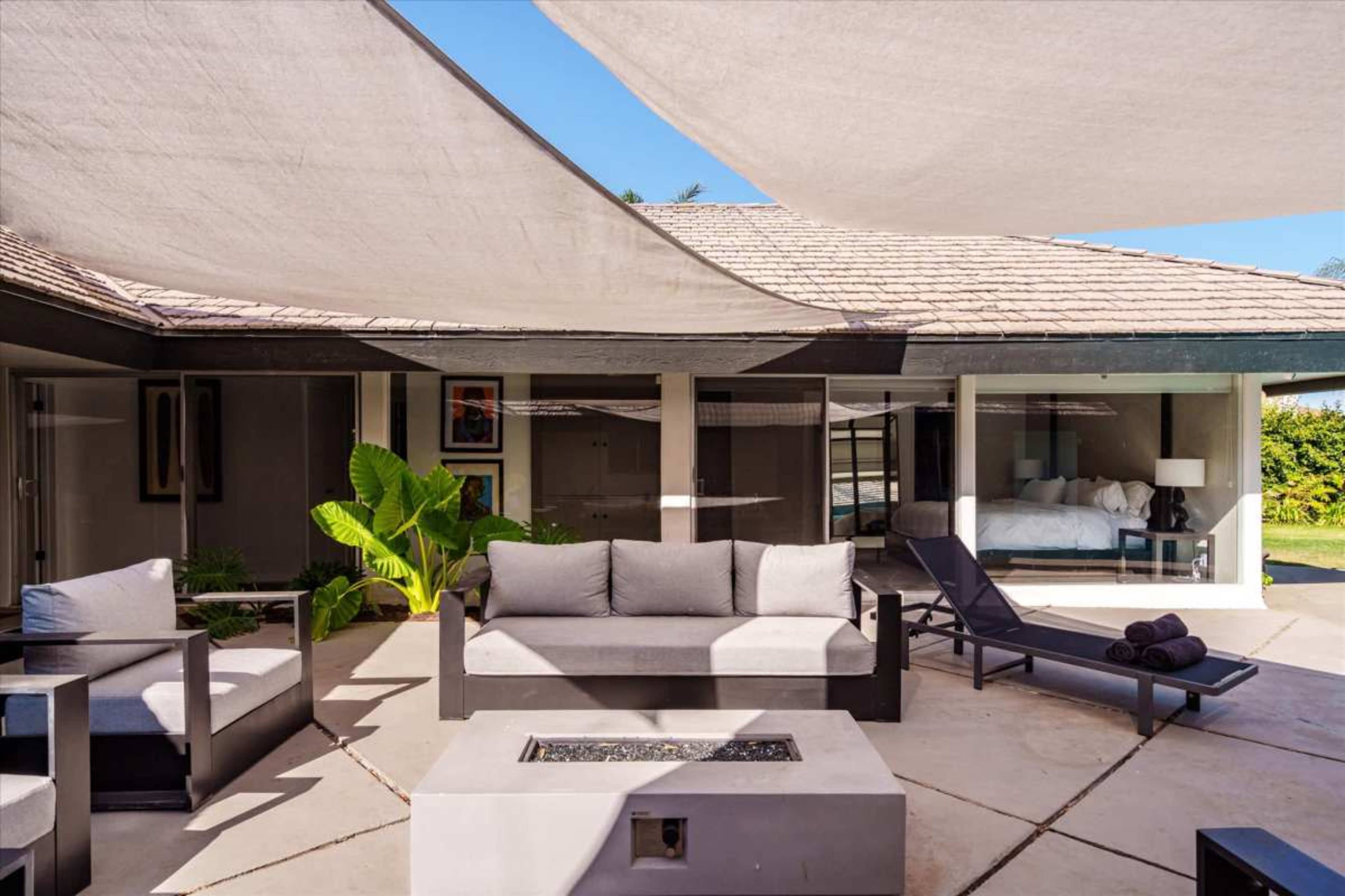 The image shows a modern outdoor patio area with a fire pit, comfortable seating, and shaded areas created by large fabric canopies.