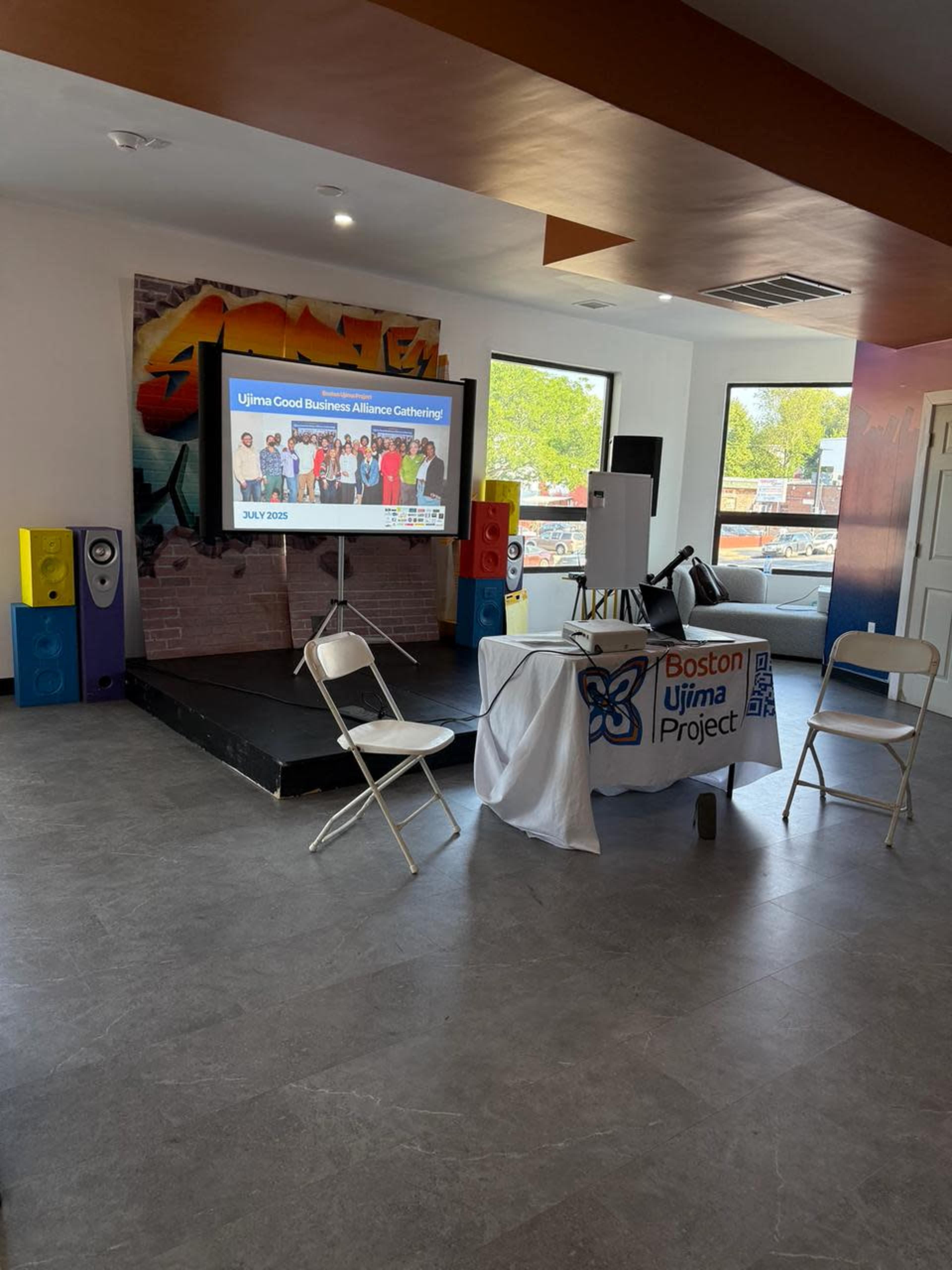 The image shows a small event space with a stage, a table displaying a sign for the "Boston Ujima Project," and several chairs set up for attendees.